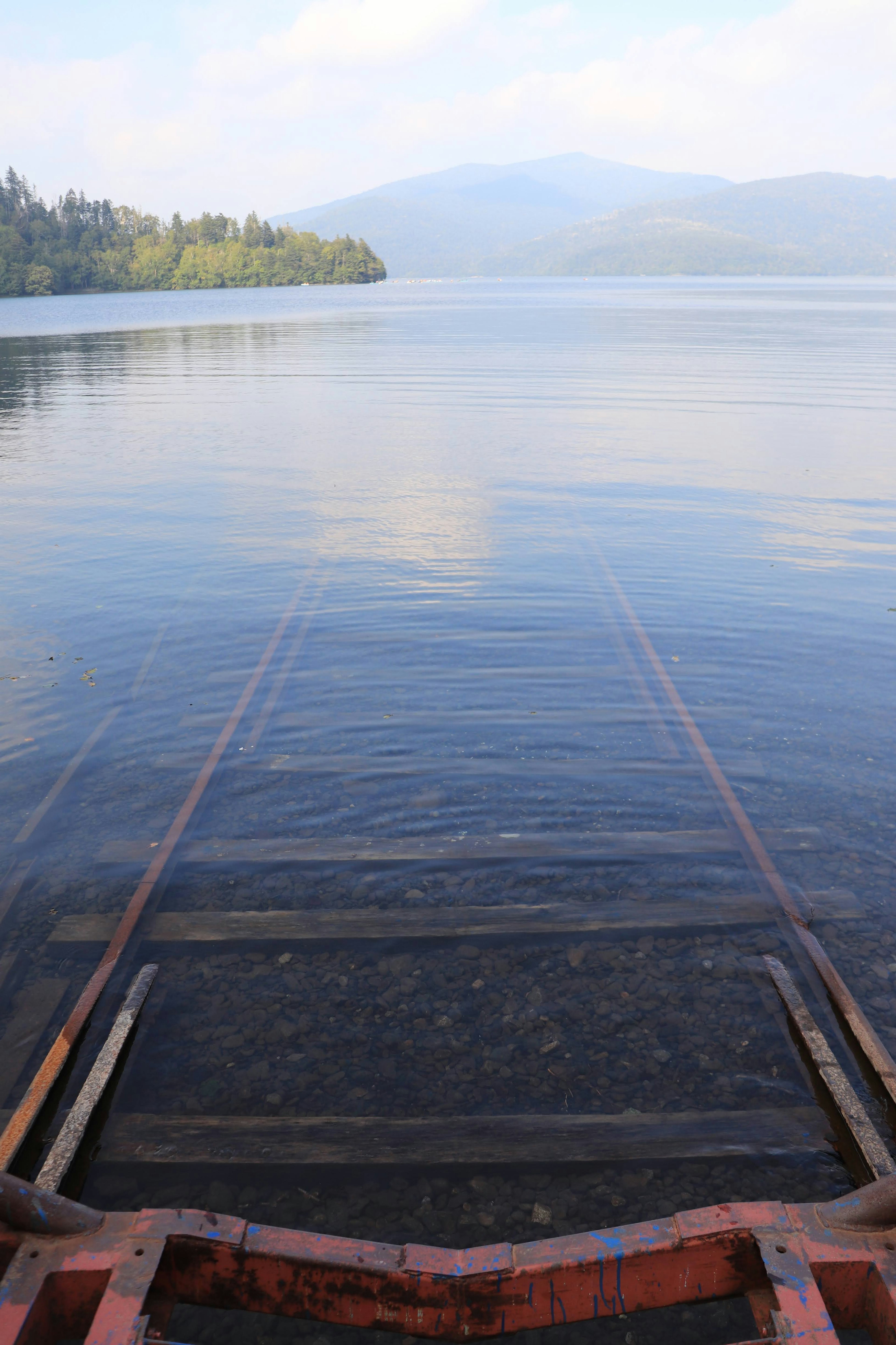 Jembatan kayu yang mengarah ke danau tenang dengan pegunungan di latar belakang