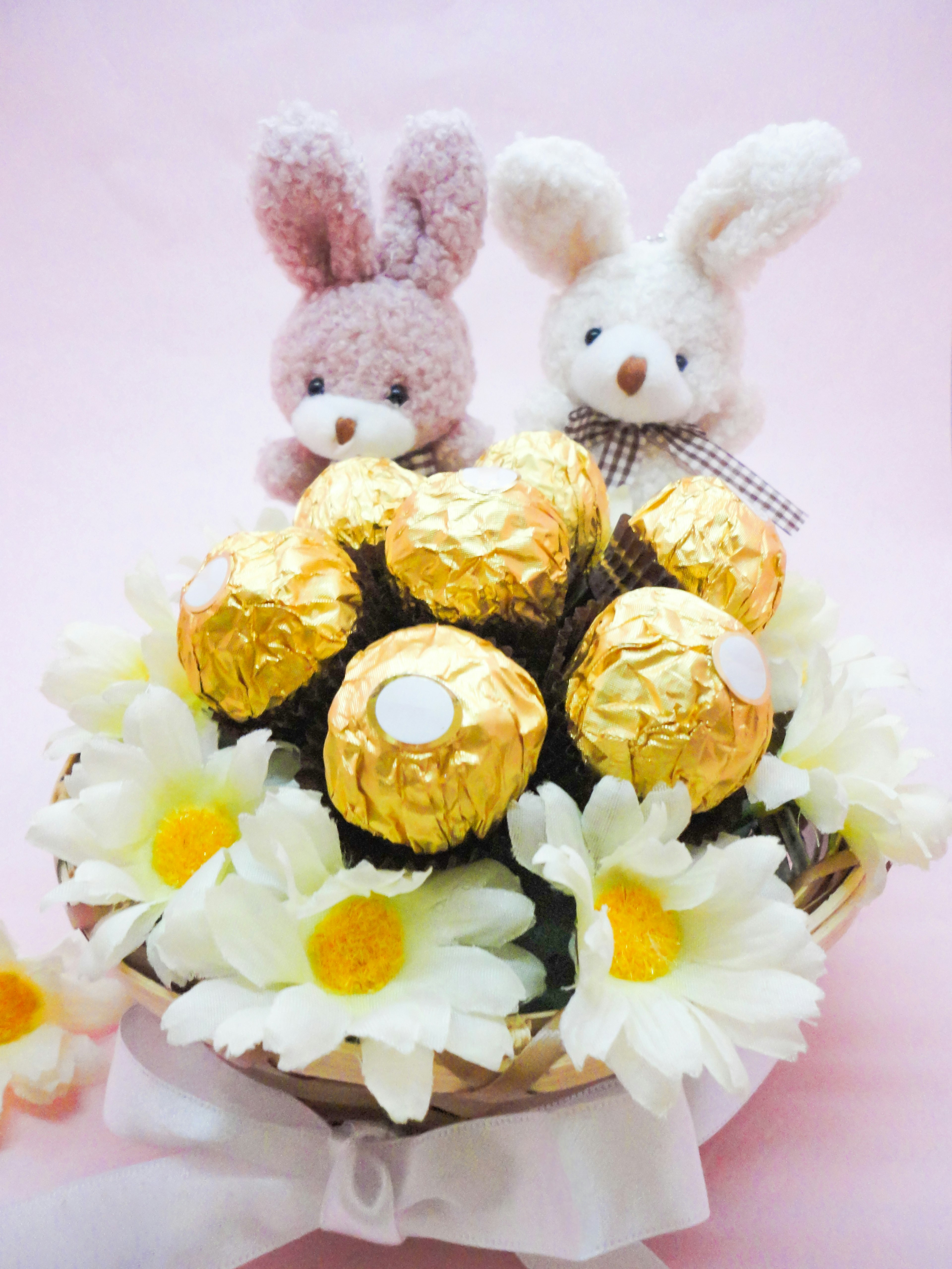 Una canasta con bolas de chocolate dorado y flores amarillas con dos juguetes de peluche de conejito sobre un fondo rosa