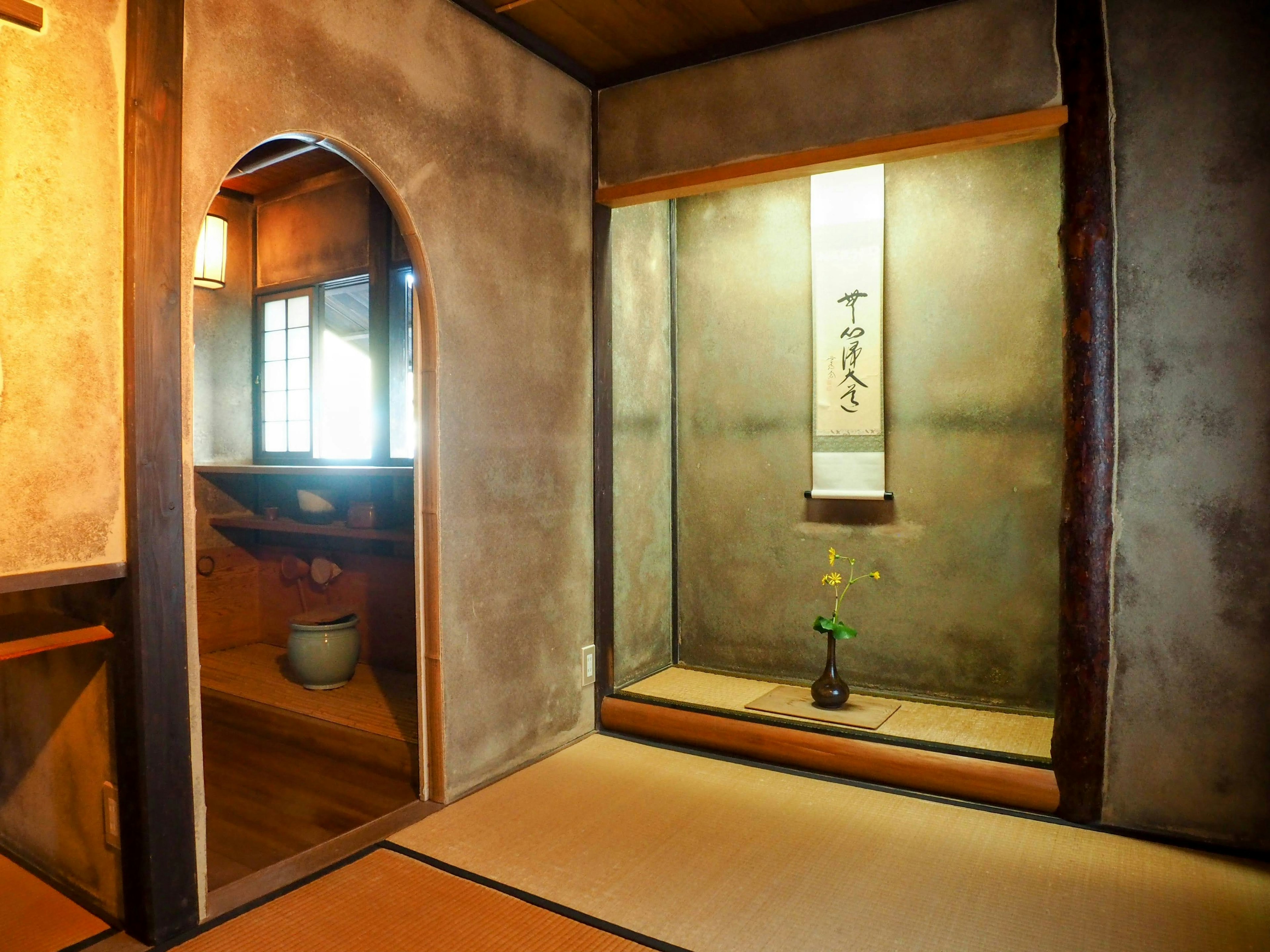 Vista interior de una habitación japonesa tradicional con un espejo arqueado a la izquierda y un rollo colgado con un arreglo floral a la derecha