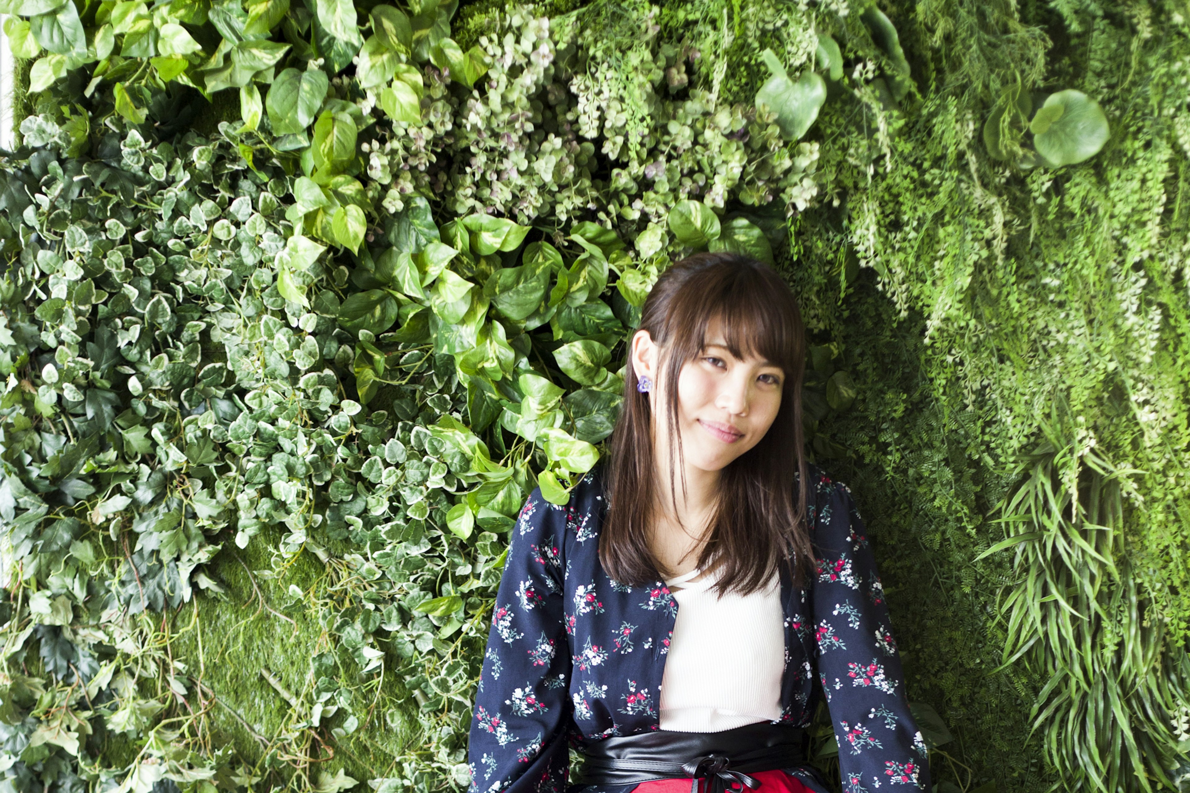 Retrato de una mujer sentada frente a una pared verde rodeada de plantas exuberantes