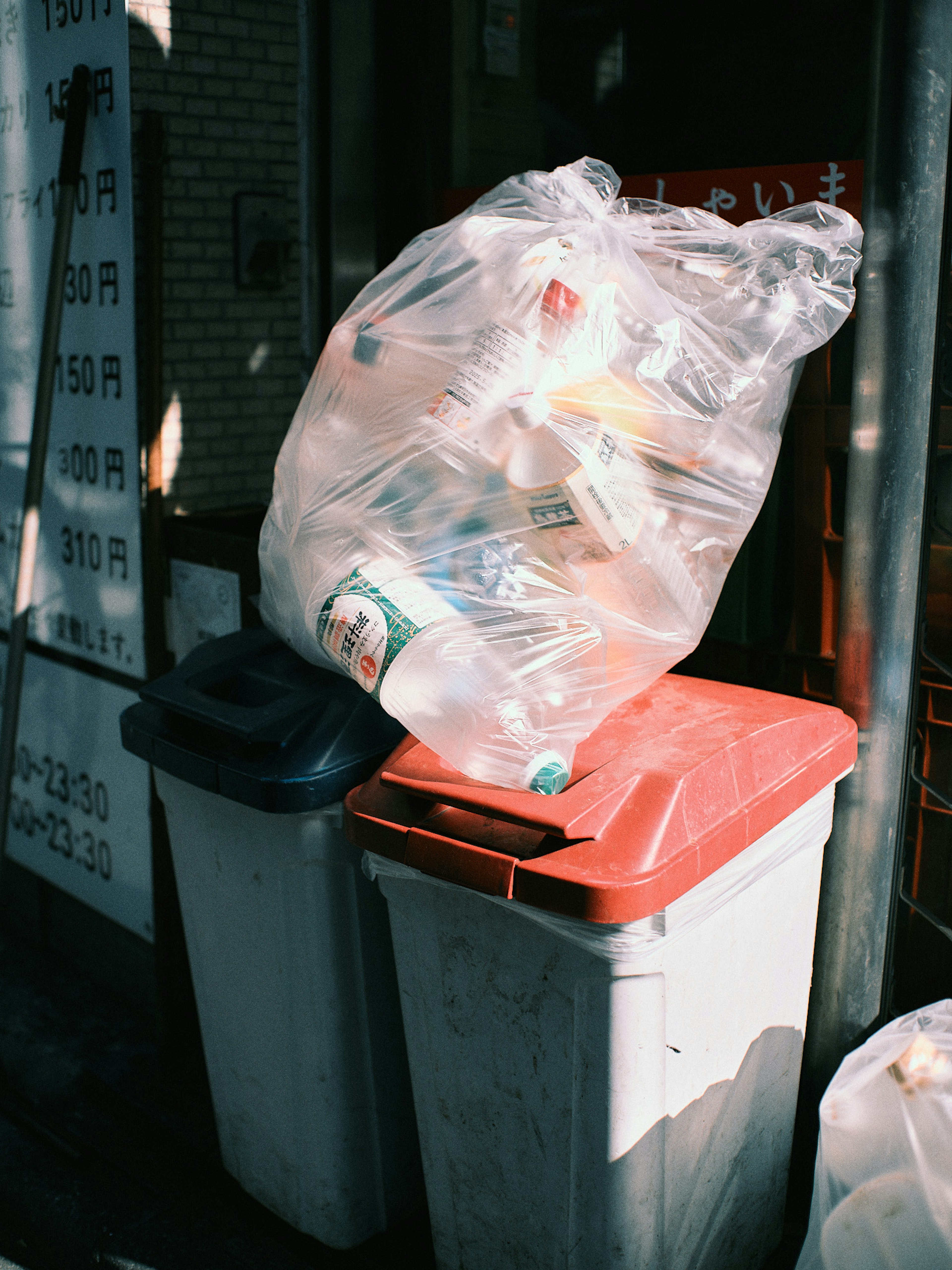 Sebuah kantong sampah transparan yang penuh dengan sampah ditumpuk di atas tempat sampah merah