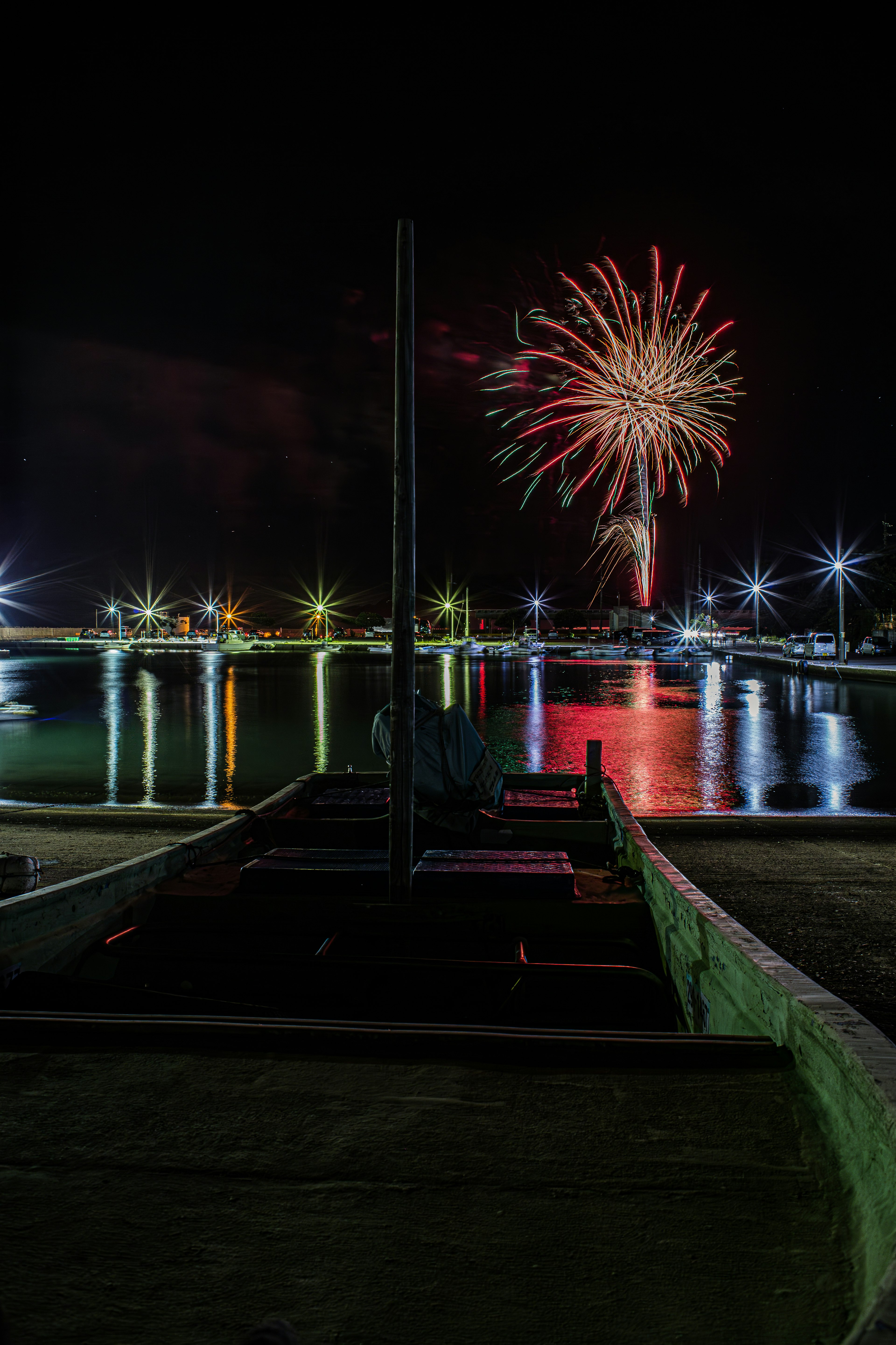 Fuegos artificiales iluminando el cielo nocturno con reflejos en el agua y un bote en primer plano