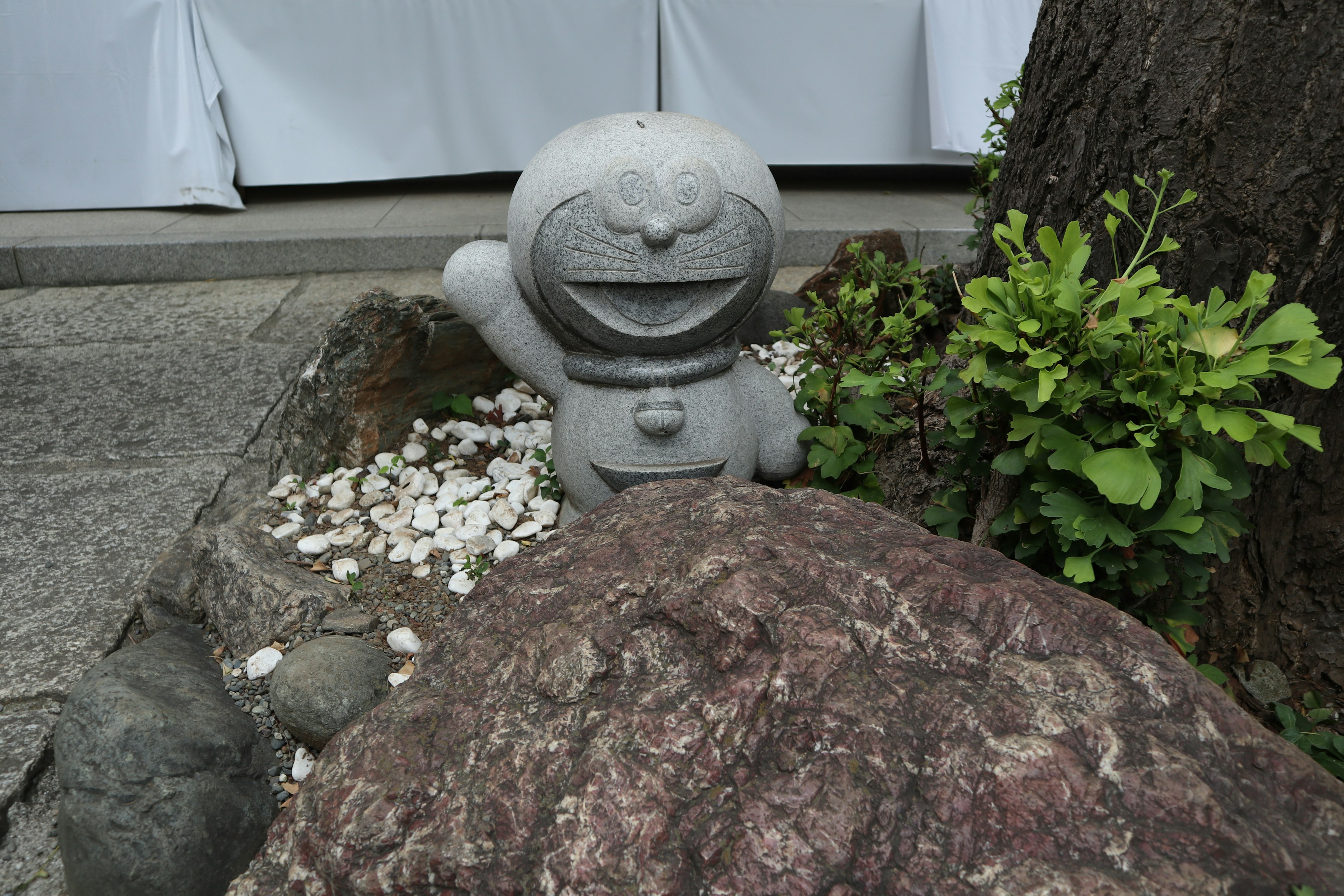 A stone statue of Doraemon standing next to a tree