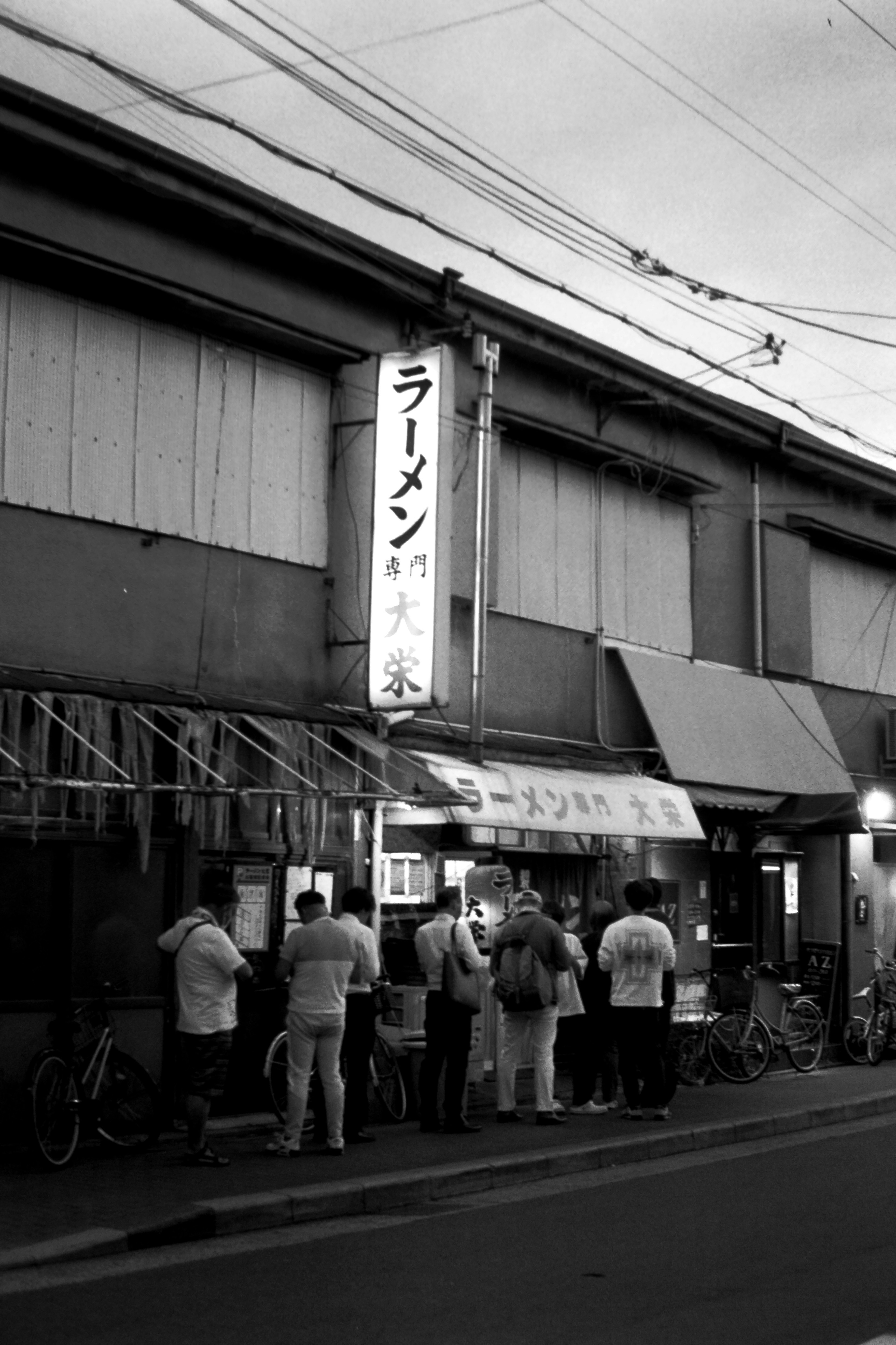 夜のラーメン屋の前に並ぶ人々と看板