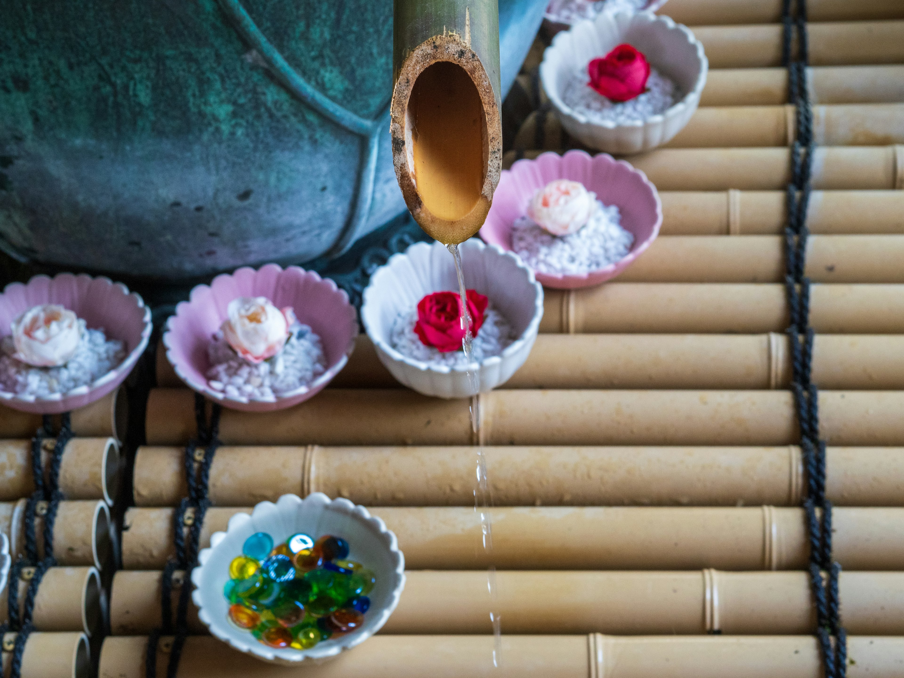 Agua fluyendo de un tubo de bambú a pequeños cuencos con pétalos de flores y cuentas coloridas sobre una estera de bambú