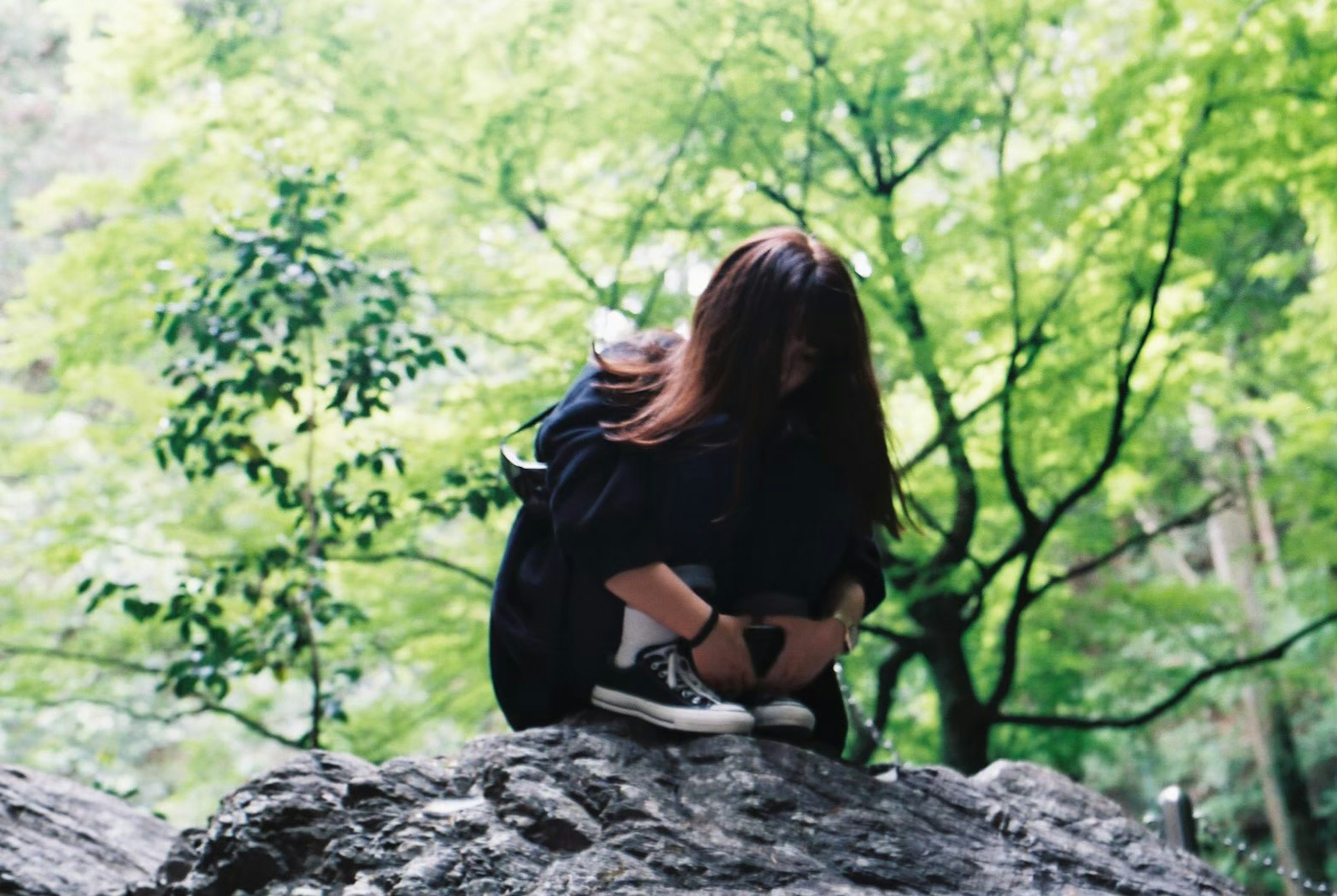 緑の木々に囲まれた岩の上に座る女性彼女は膝を抱えた姿勢でいる