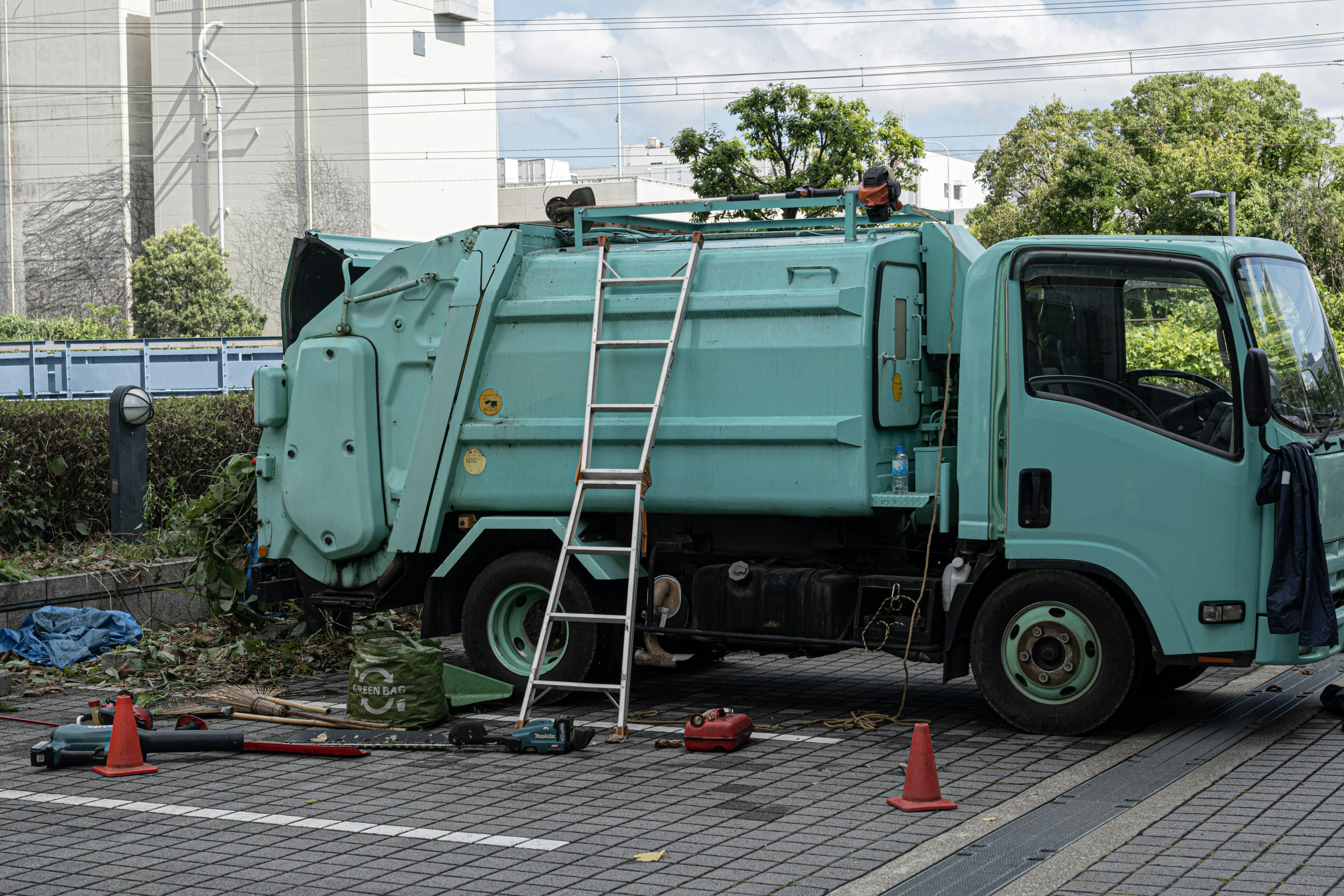 Un camion della spazzatura verde parcheggiato in un'area con una scala e attrezzi nelle vicinanze circondati da piante