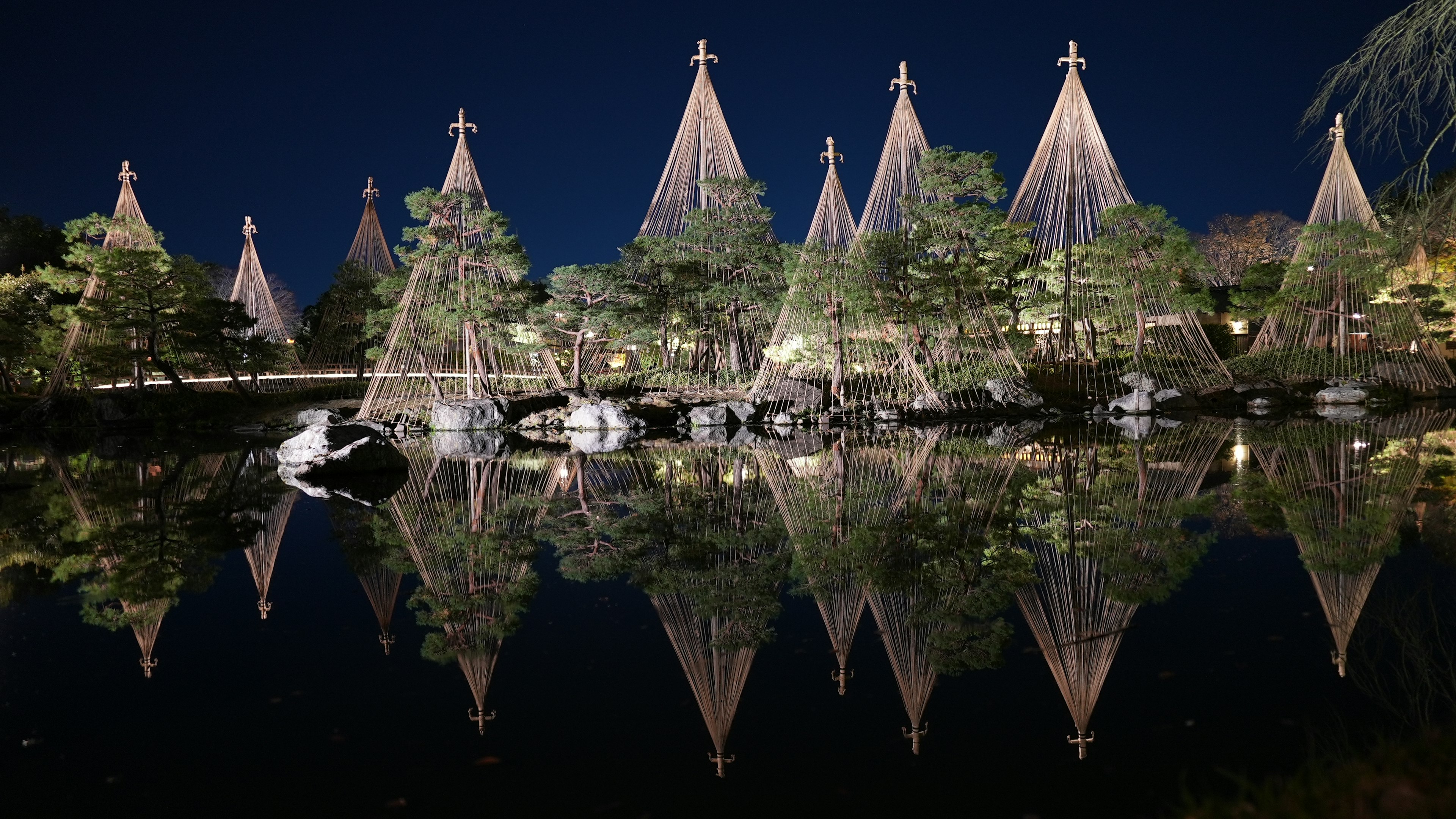 Schöner japanischer Garten mit Kiefern und traditionellen Bambusstützen, die nachts in einem Teich reflektiert werden