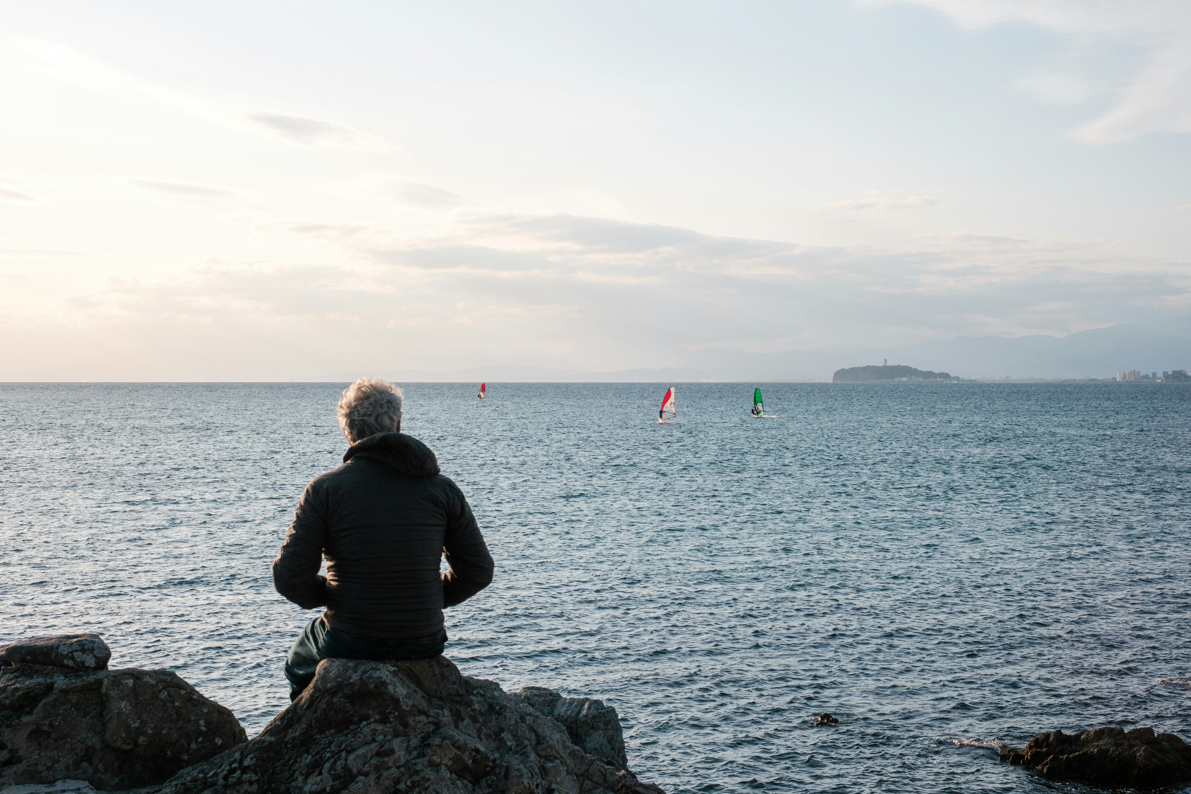 Mann sitzt auf einem Stein am Meer mit Segelboards im Hintergrund