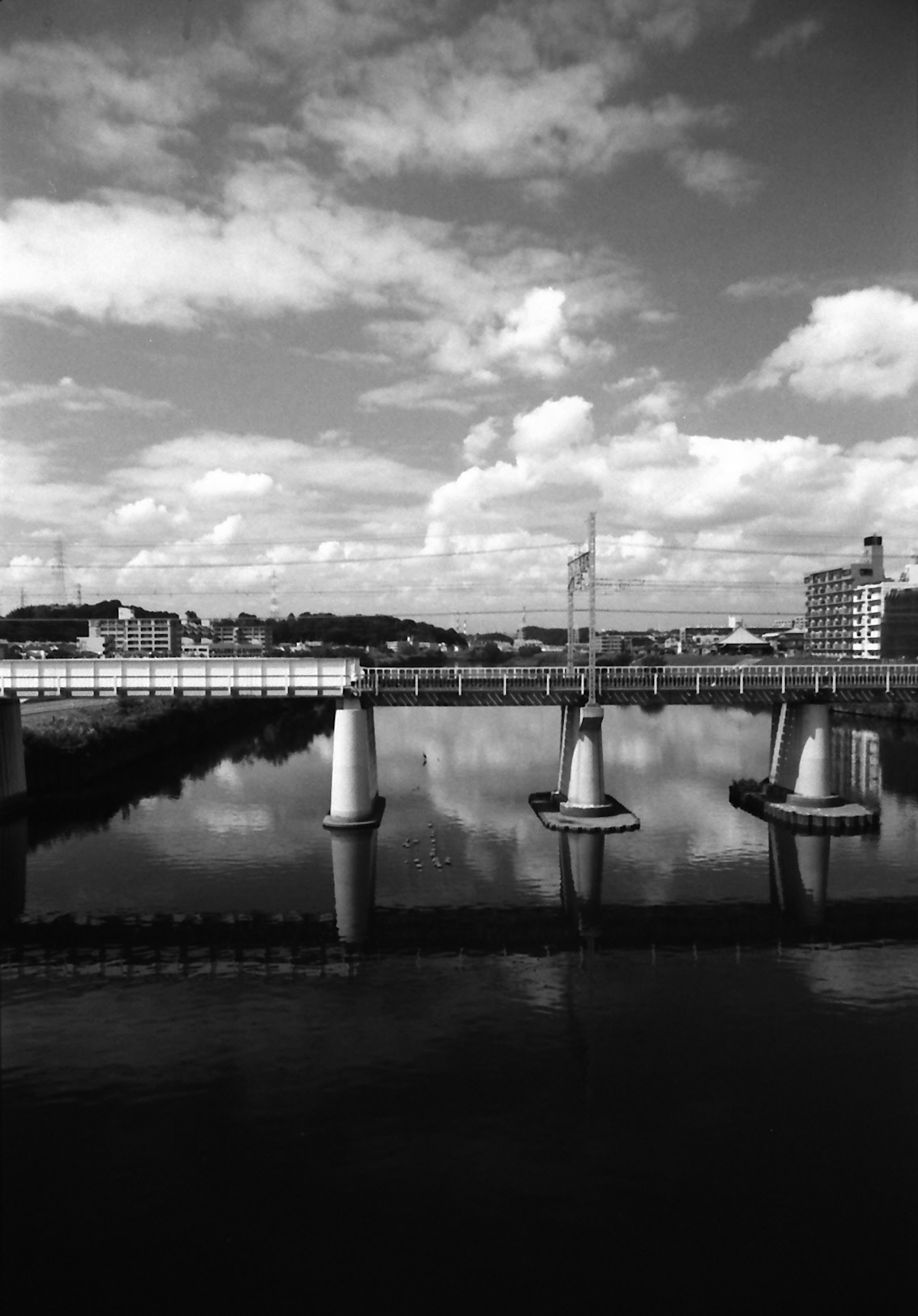 Foto de paisaje en blanco y negro con agua tranquila reflejando nubes y edificios con pilares de puente