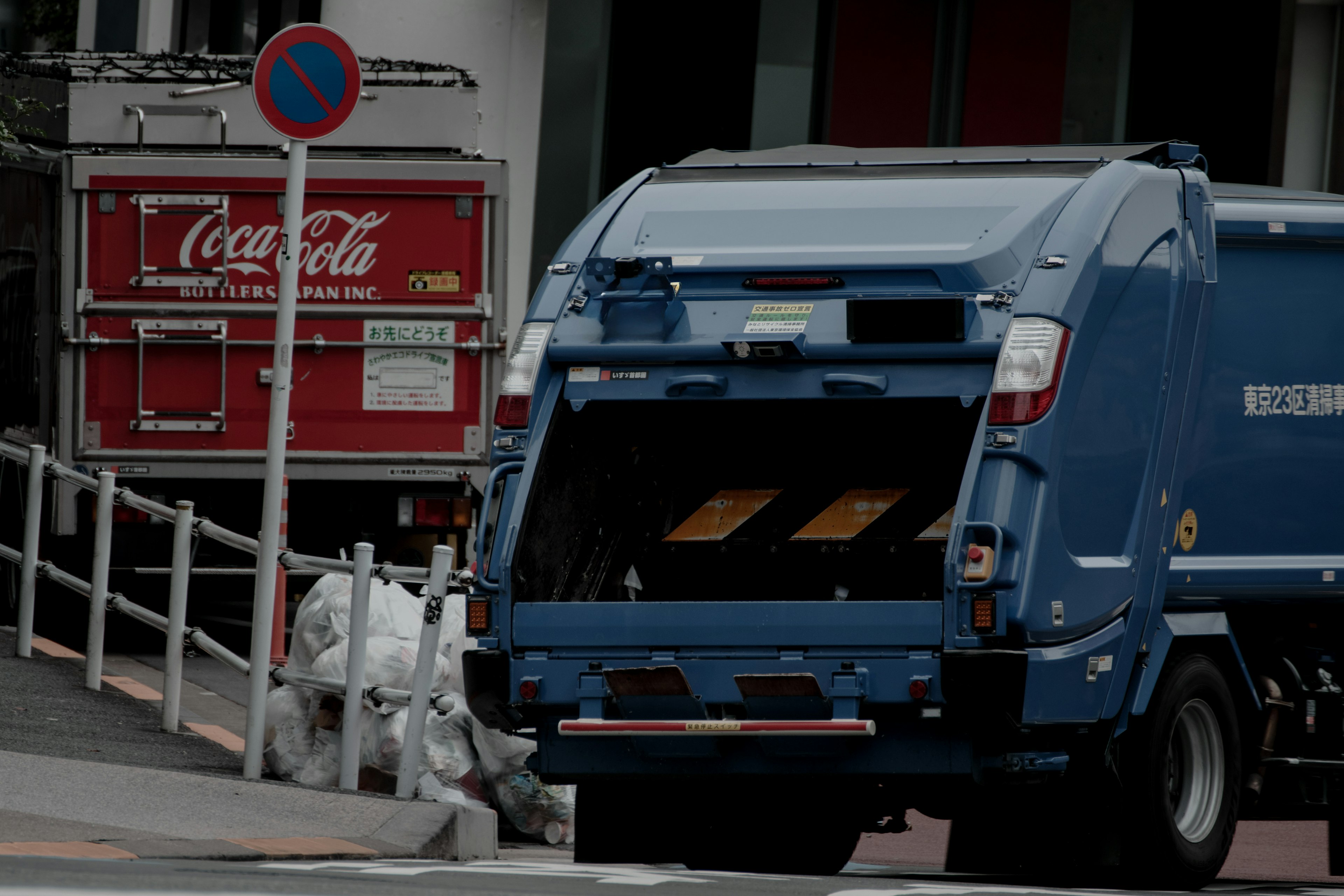 Blue garbage truck parked with its back open showing waste inside Red Coca-Cola sign in the background