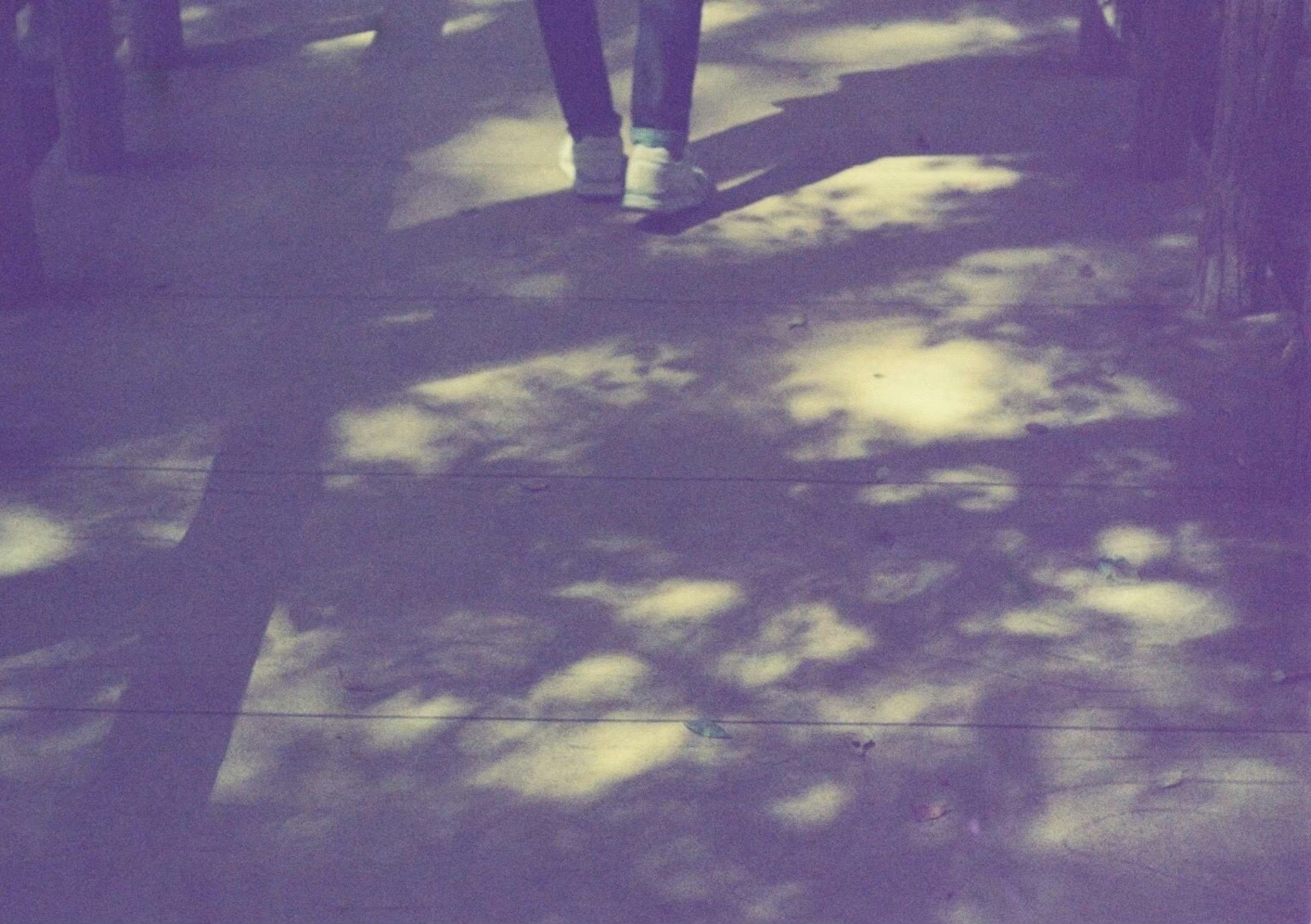 A person's feet walking on a sidewalk with scattered shadows