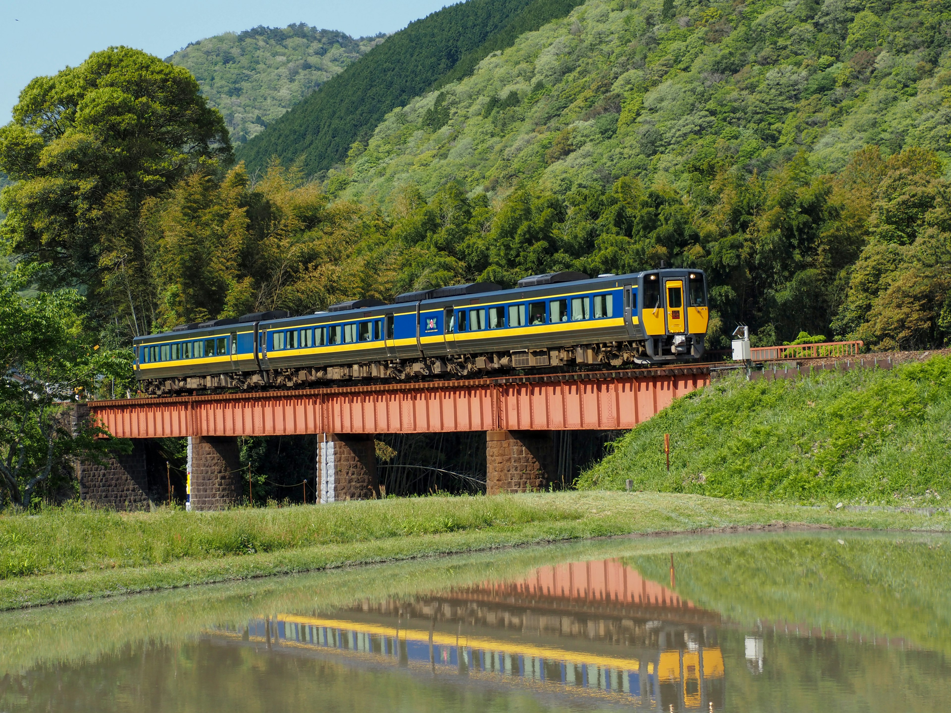 Un train bleu et jaune traversant un pont rouge avec des montagnes verdoyantes en arrière-plan