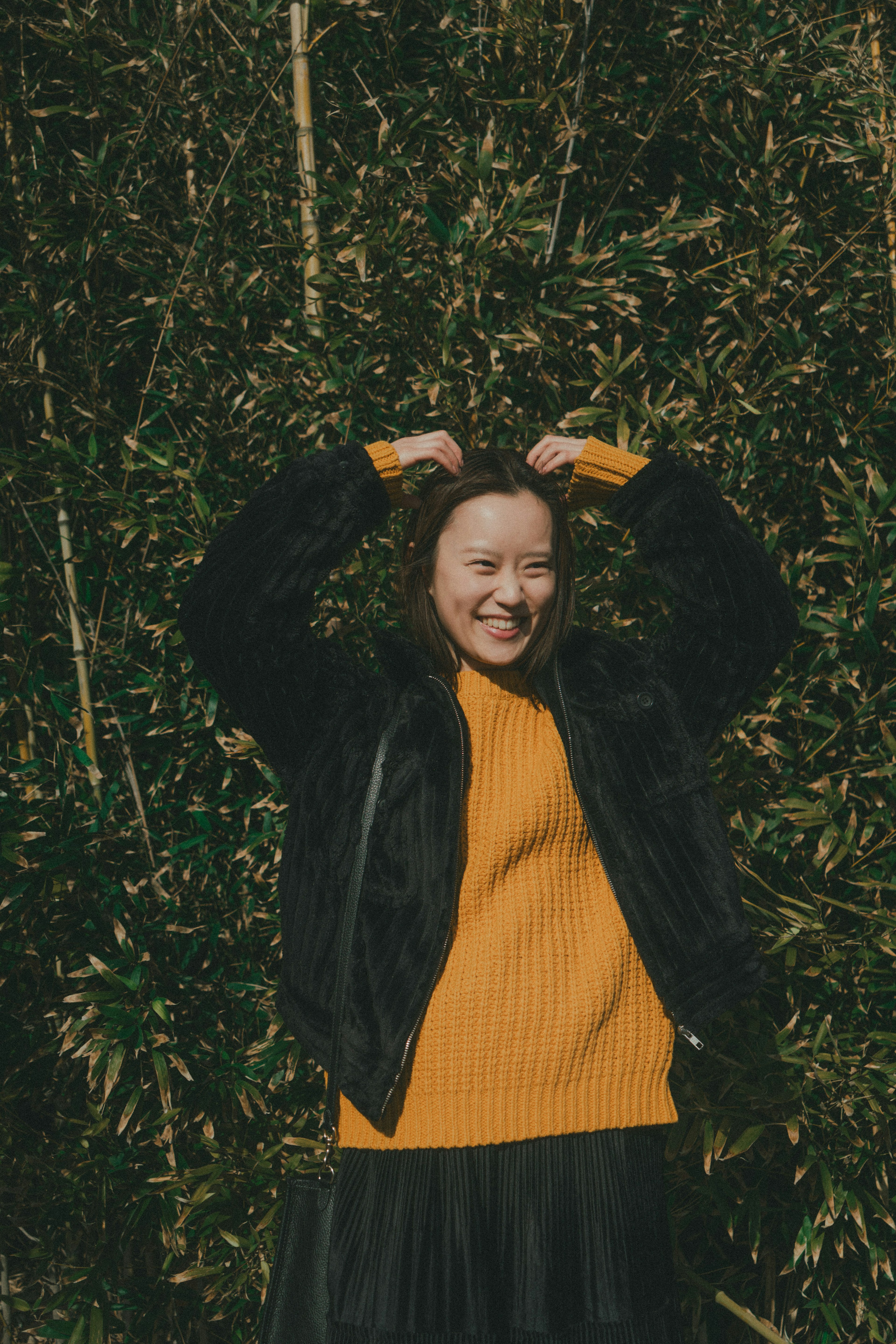 Une femme en veste noire et pull orange souriante et posant devant un feuillage vert