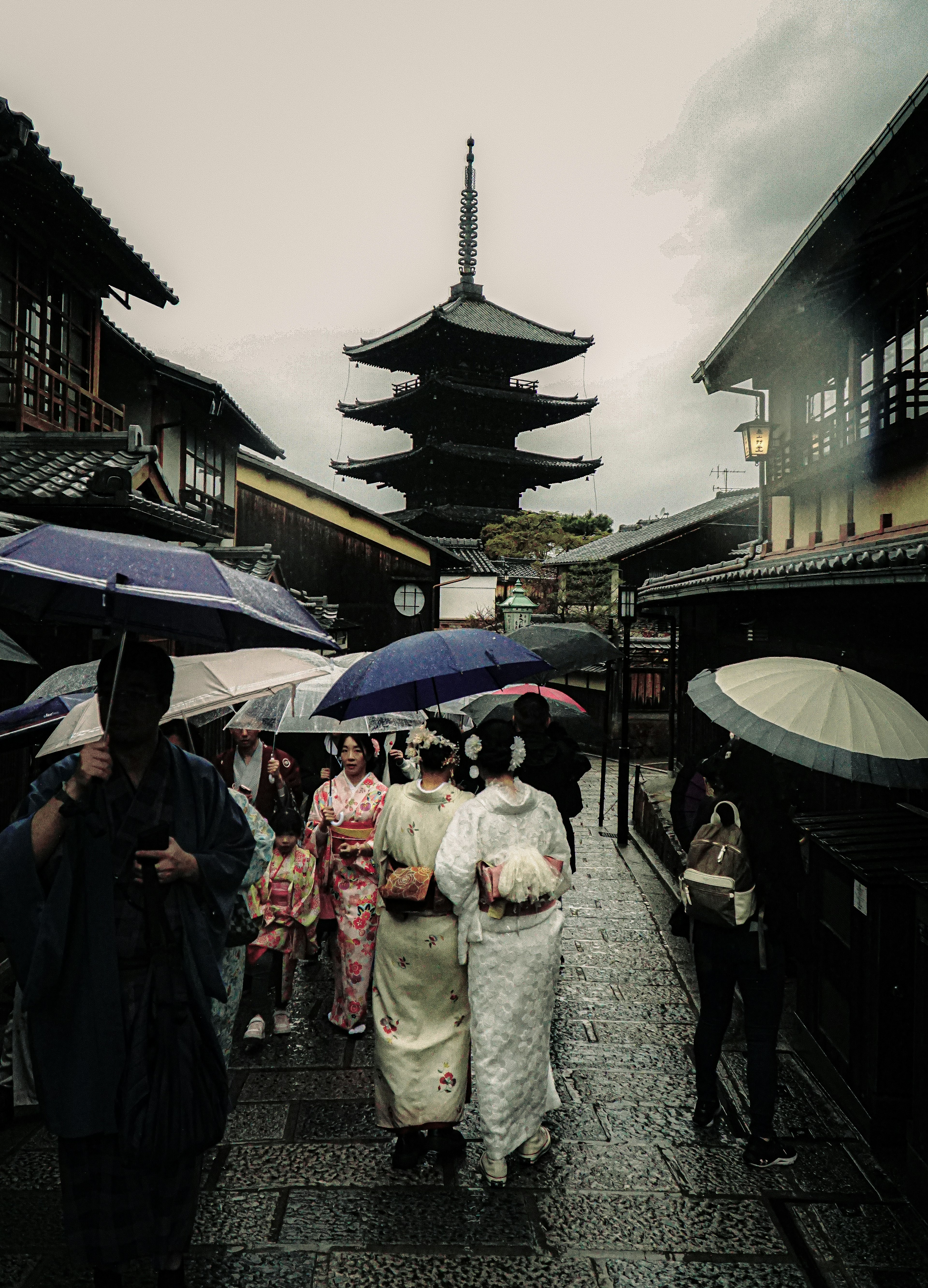 雨の中、傘を持った着物姿の人々が古い街並みを歩いている