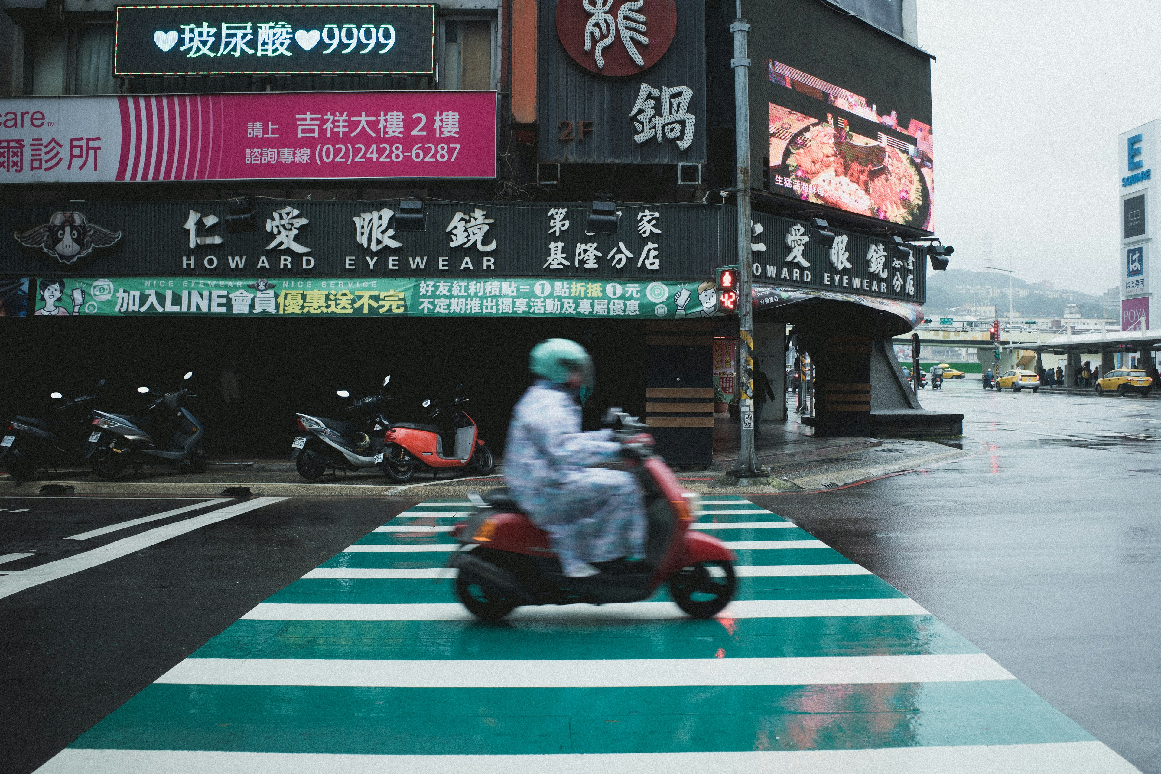 雨の日の交差点で走行する赤いスクーターと緑の横断歩道の風景