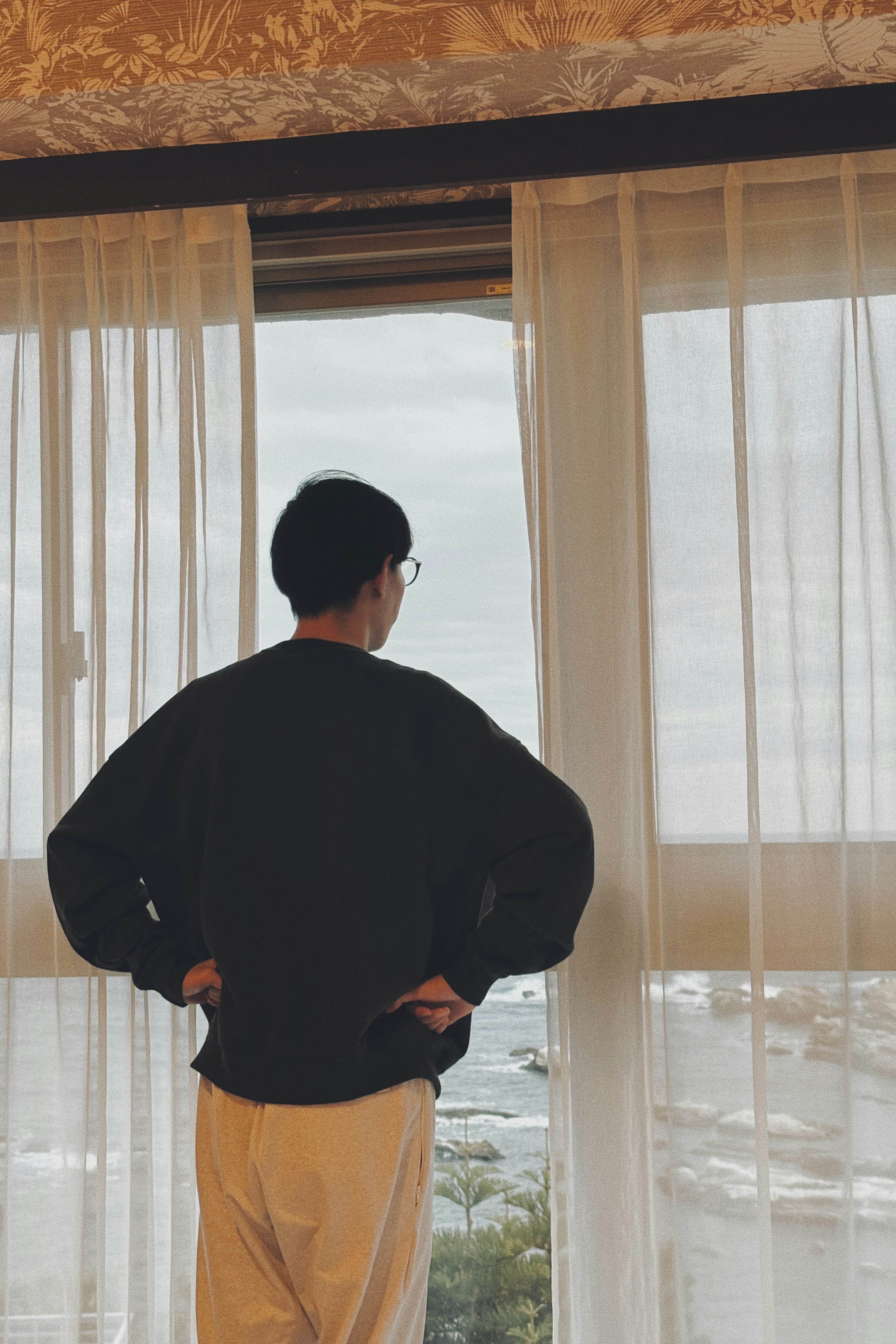 A man standing with his back to the window looking out curtains are drawn