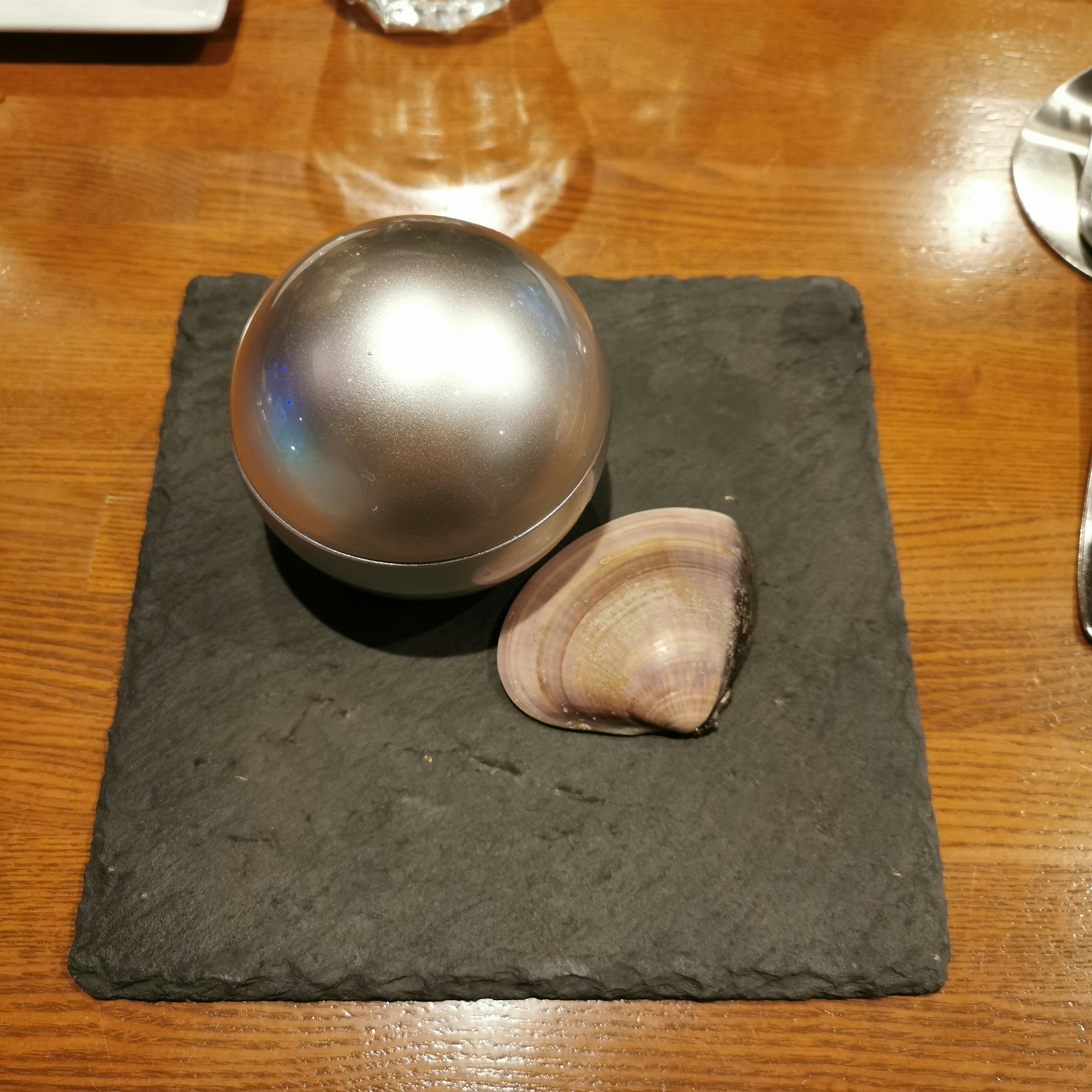 Un objet sphérique argenté et une coquille de palourde placés sur une assiette en pierre noire