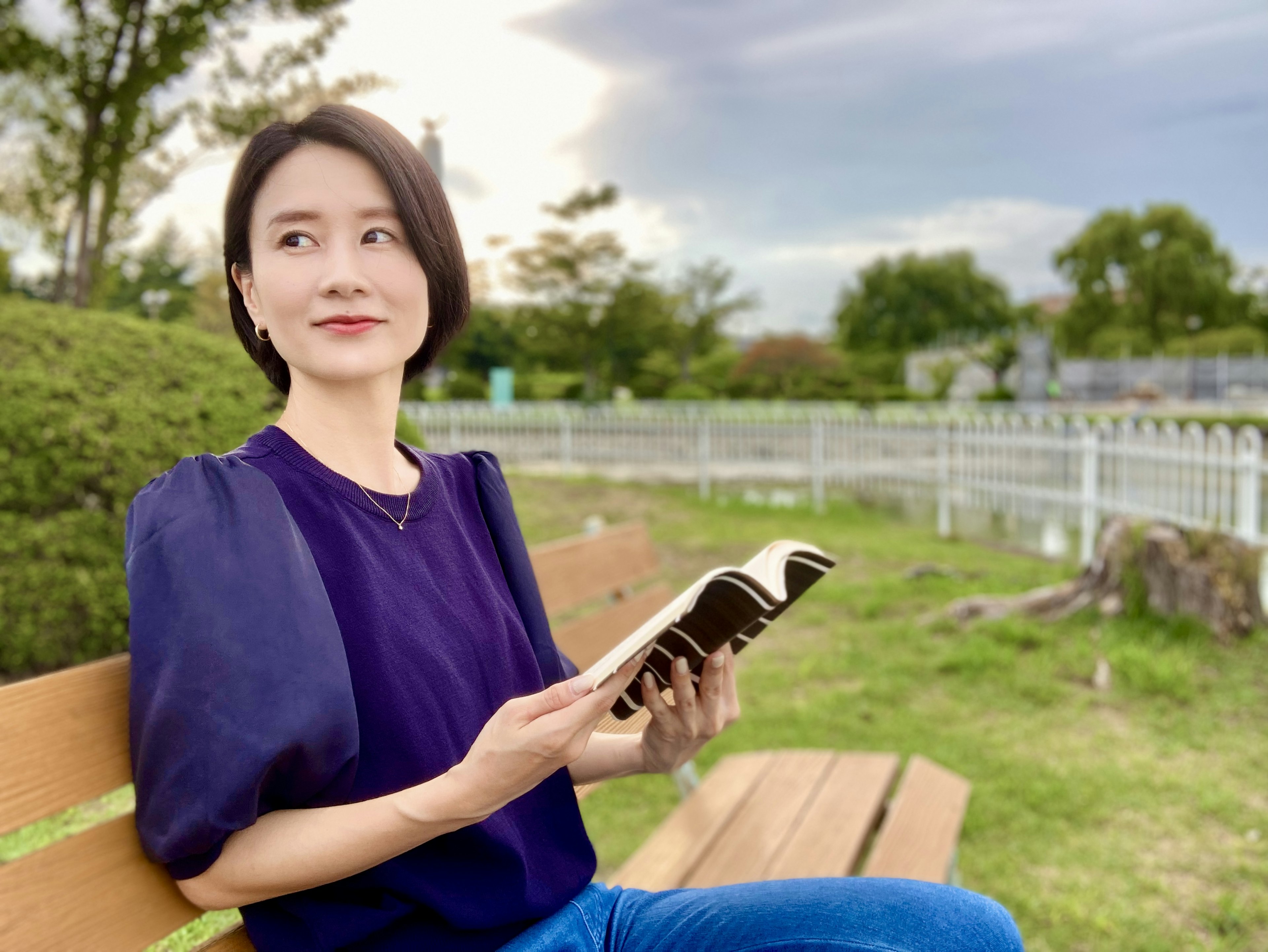 Porträt einer Frau, die ein Buch in einem Park hält, mit grünem Hintergrund und einem ruhigen Himmel