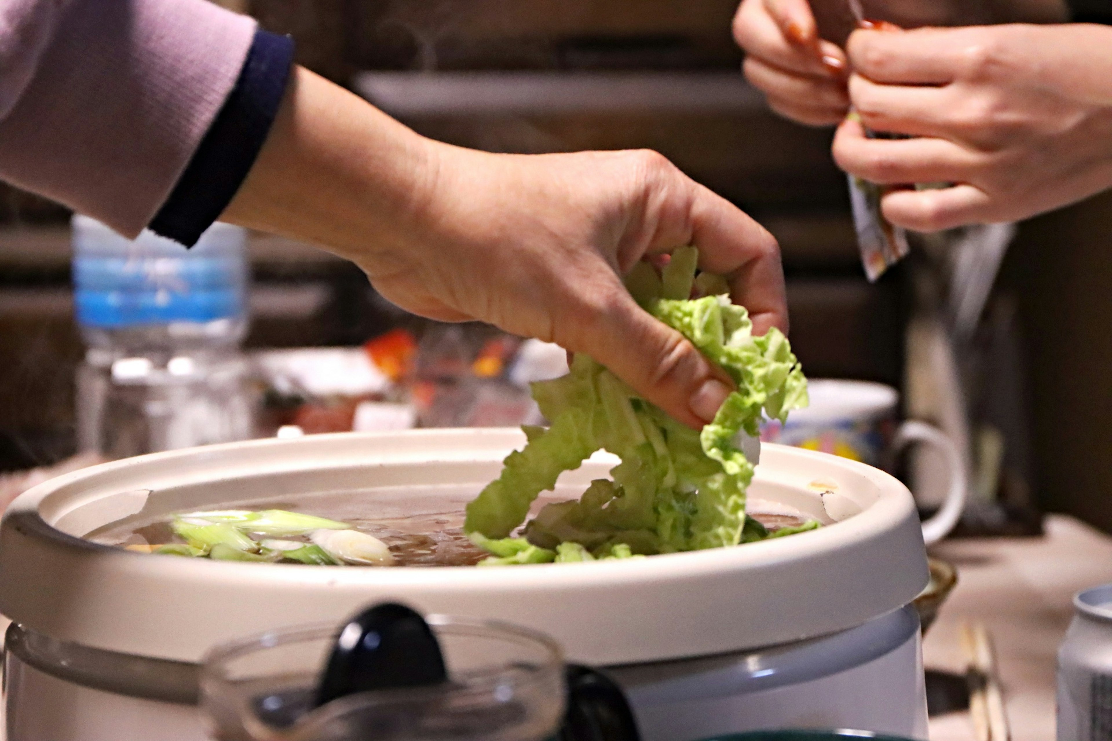 Manos agregando lechuga a una olla caliente con otra mano preparando ingredientes