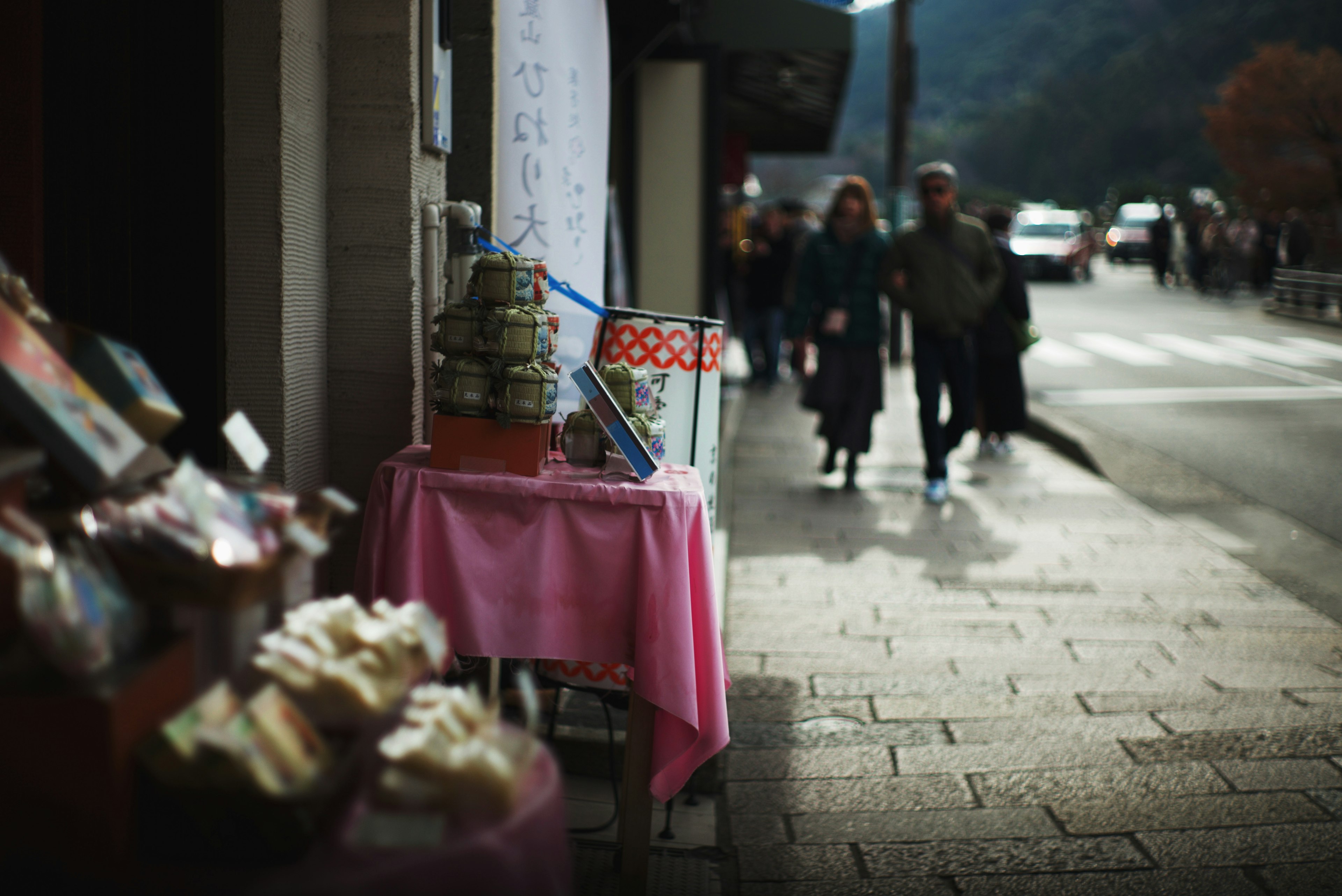 通りに面した小さな店の前にあるピンクのテーブルと商品たちを背景に歩く人々