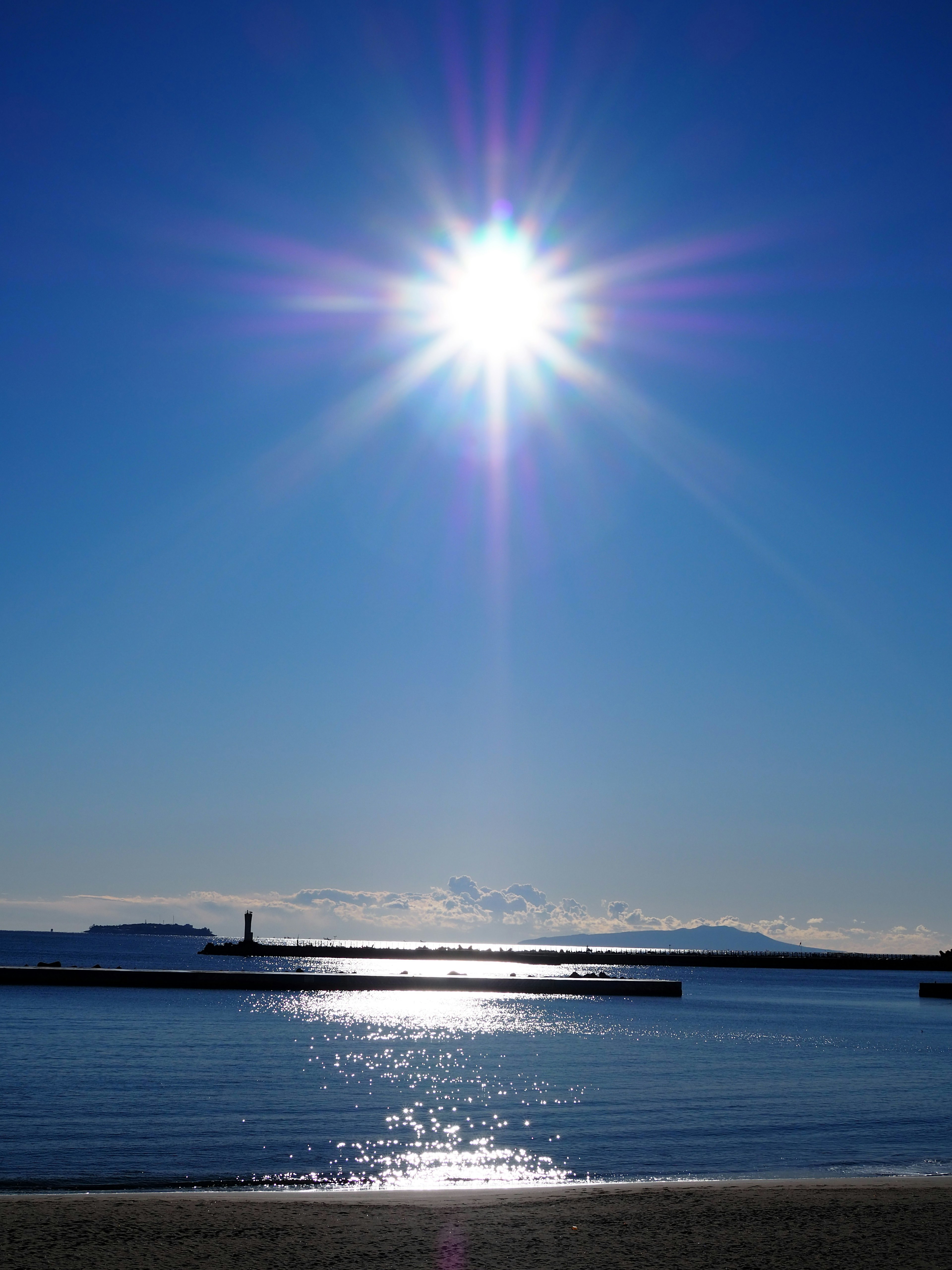 青空の下で輝く太陽の光が海に反射し、穏やかな波の様子が見える風景