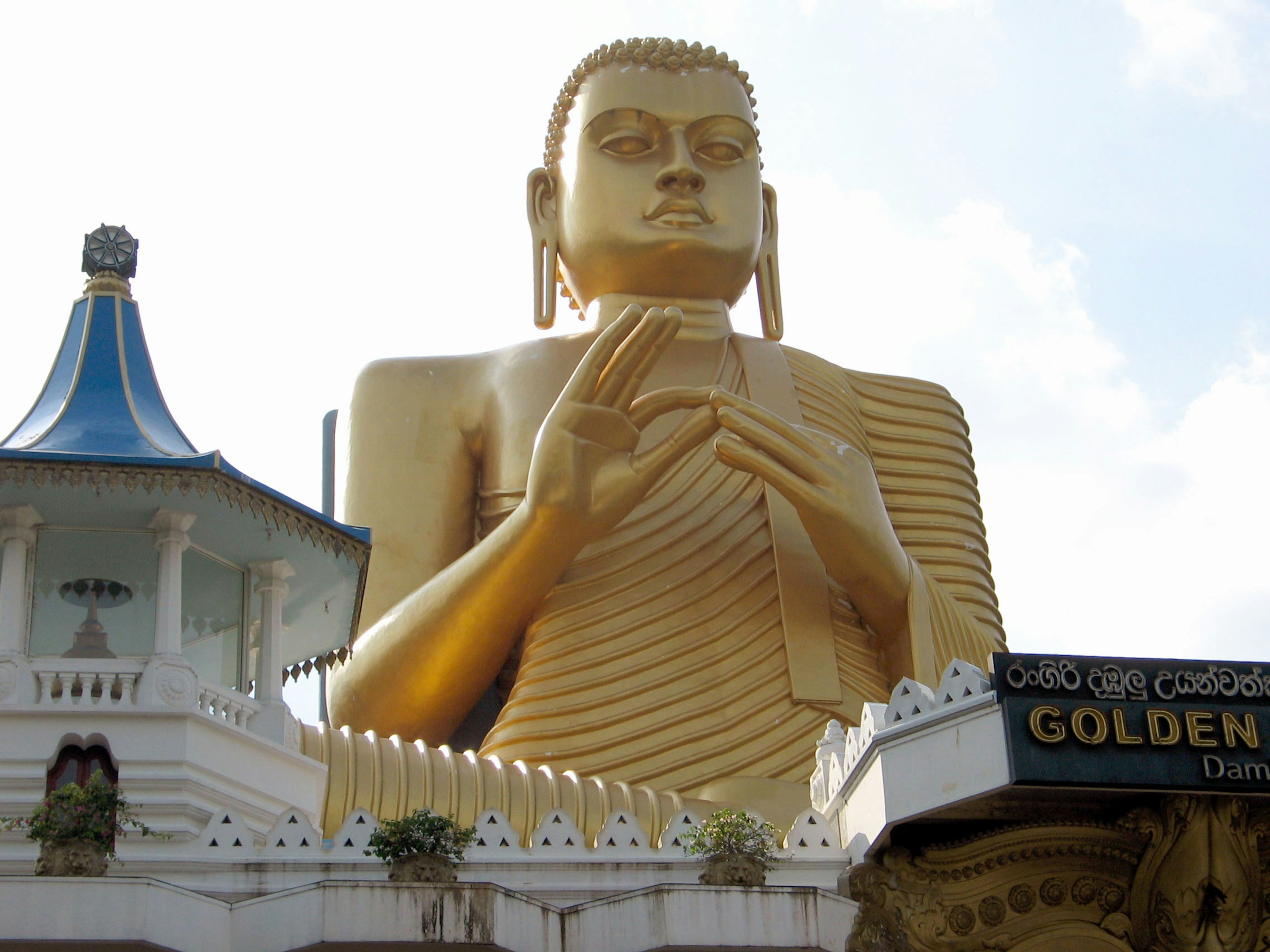 พระพุทธรูปทองขนาดใหญ่ยืนอยู่ข้างอาคารหลังคาสีน้ำเงิน