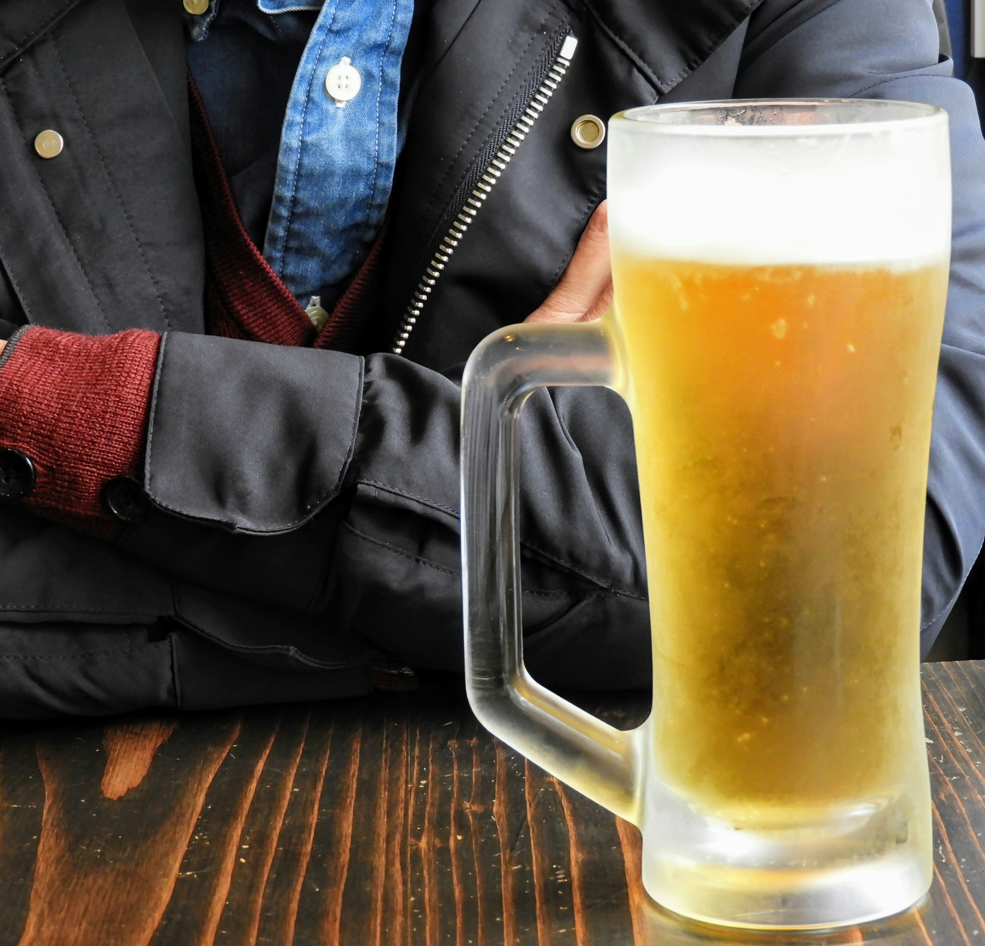 A close-up of a glass of beer on a wooden table with a person's arm in the background