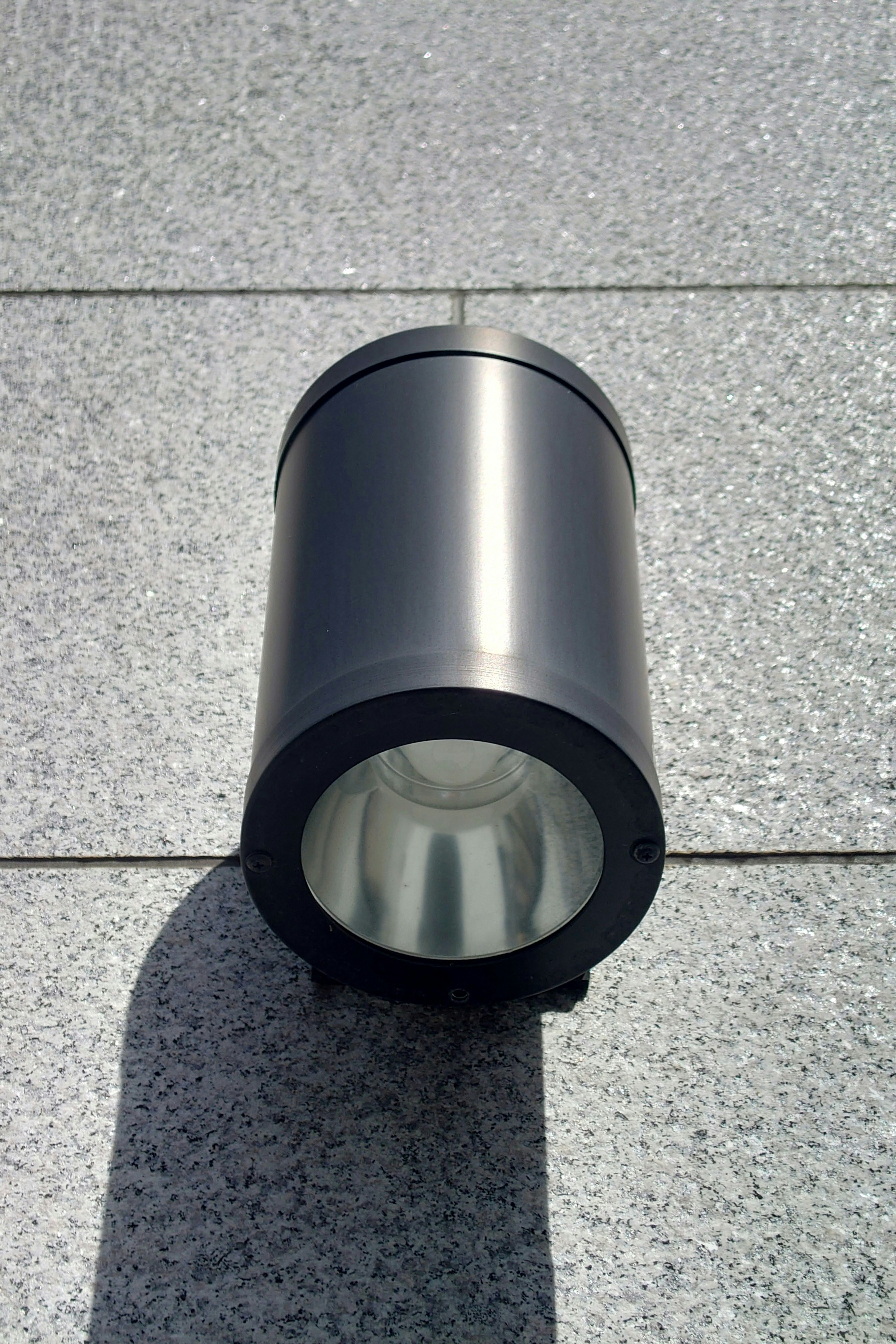 A black cylindrical lighting fixture placed on a stone floor outdoors