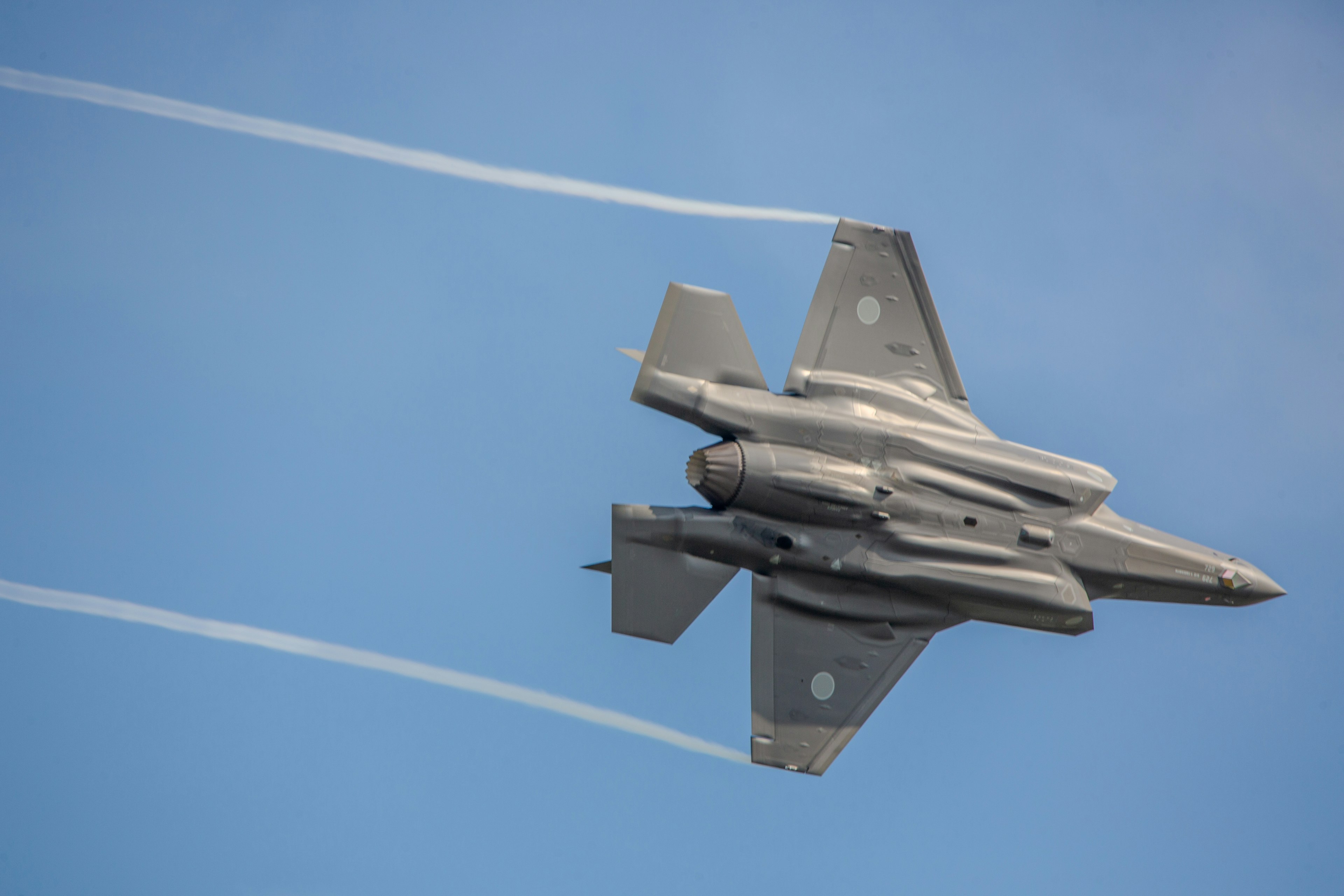 A fighter jet soaring in the blue sky with a gray body and leaving white smoke trails