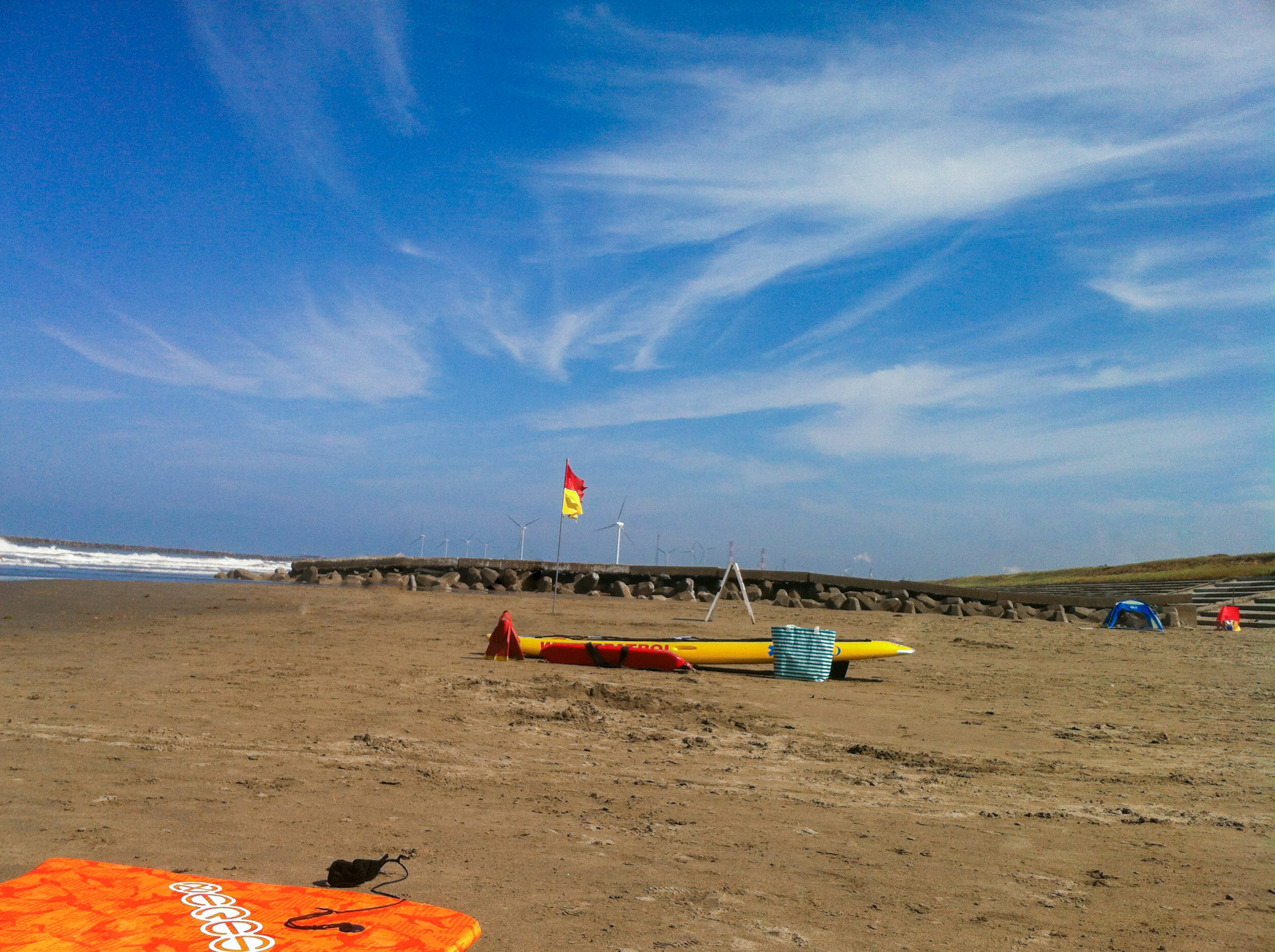 ทิวทัศน์ชายหาดใต้ท้องฟ้าสีฟ้าพร้อมเมฆสีขาว มีบอร์ดเซิร์ฟและธงช่วยชีวิตที่มองเห็นได้