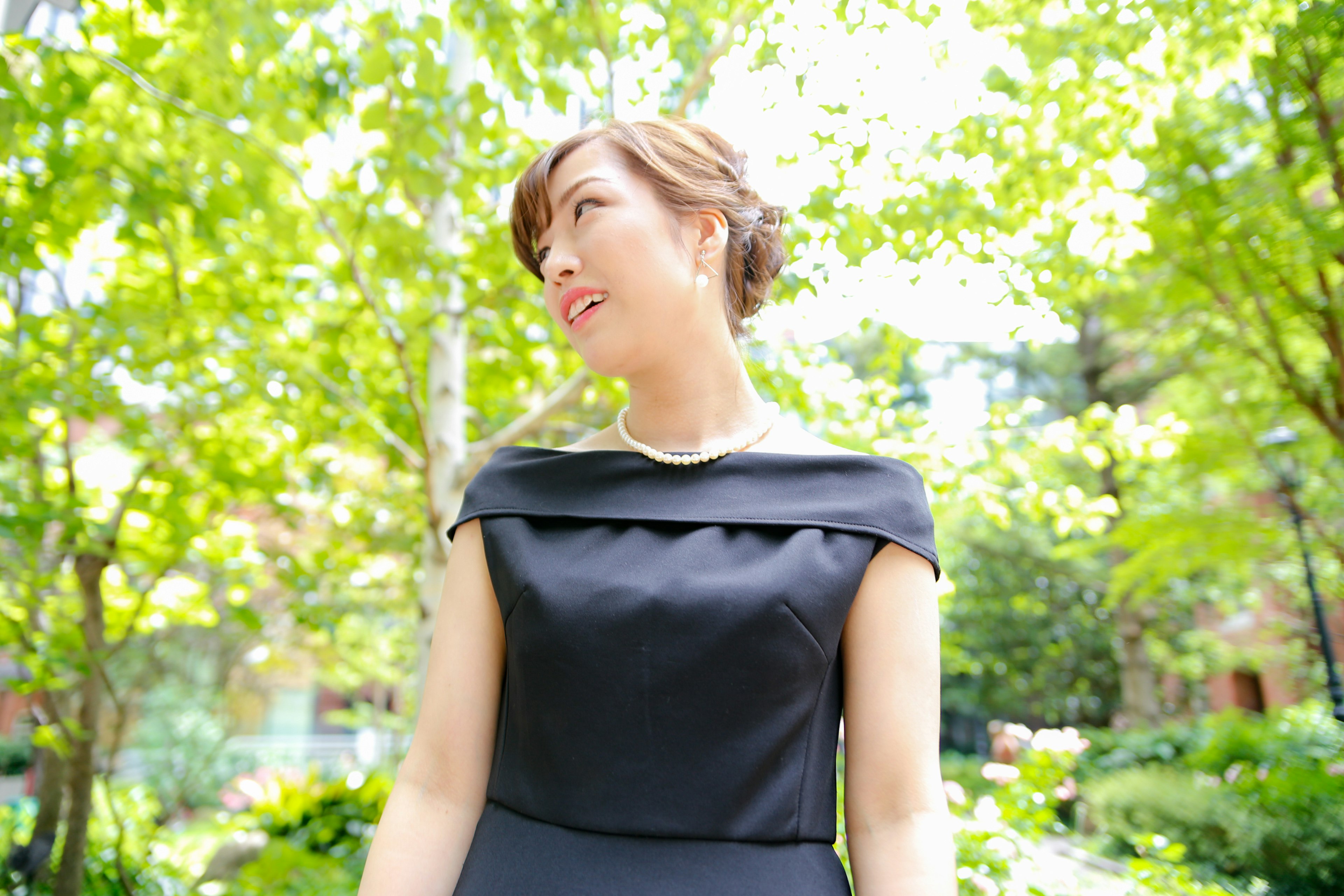 A woman in profile standing among green trees wearing a black dress