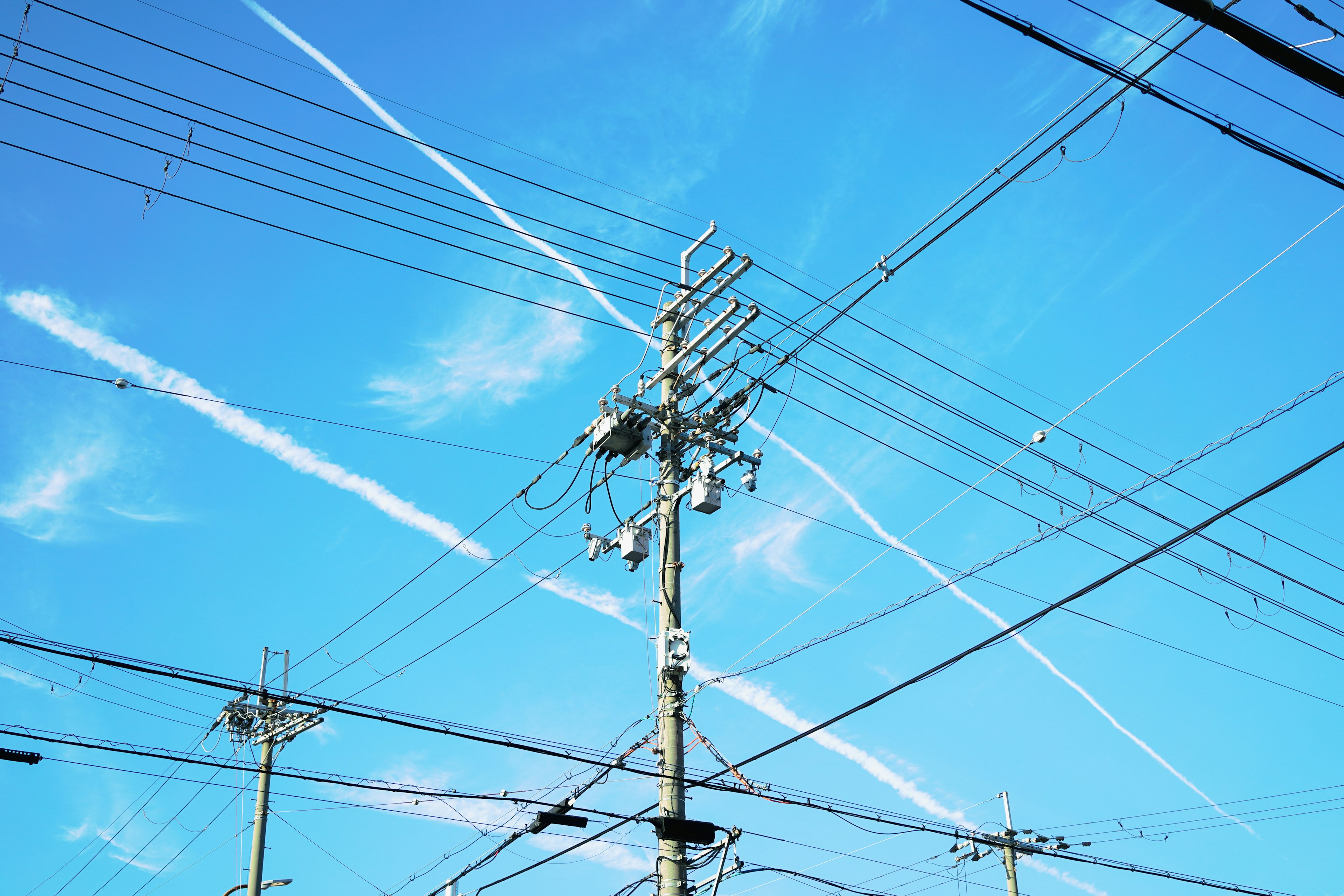 Cables eléctricos y un poste bajo un cielo azul claro con estelas de condensación cruzadas