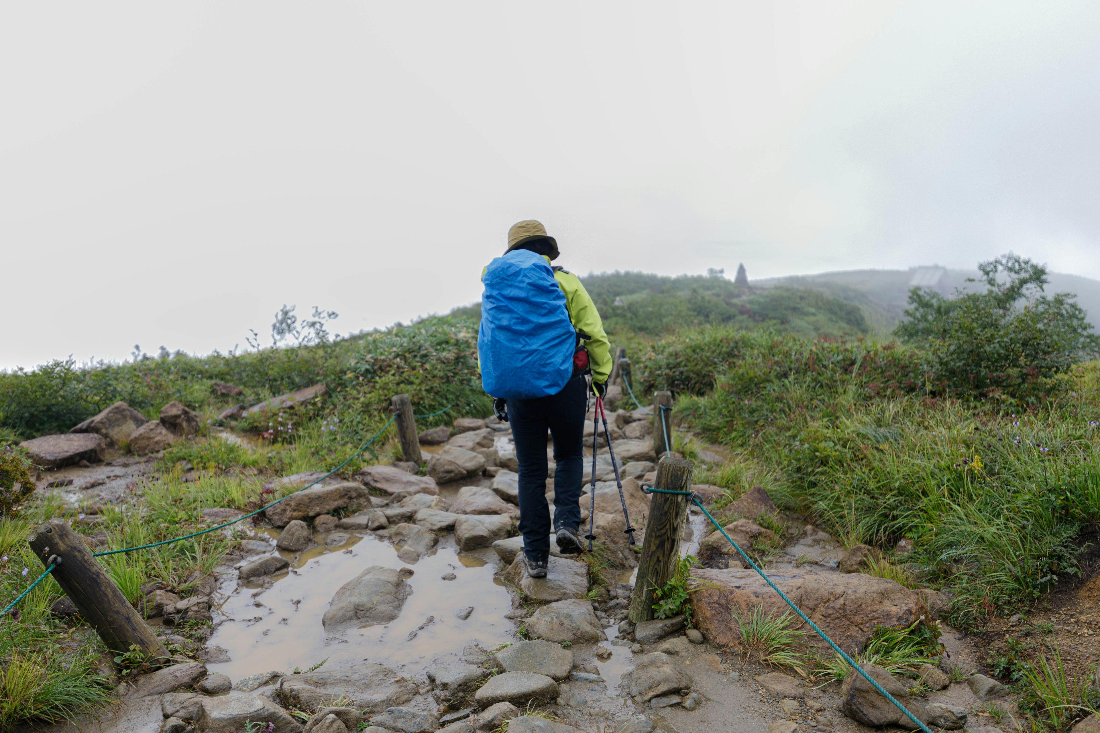 Hiker with a blue backpack walking on a rocky path in foggy mountains