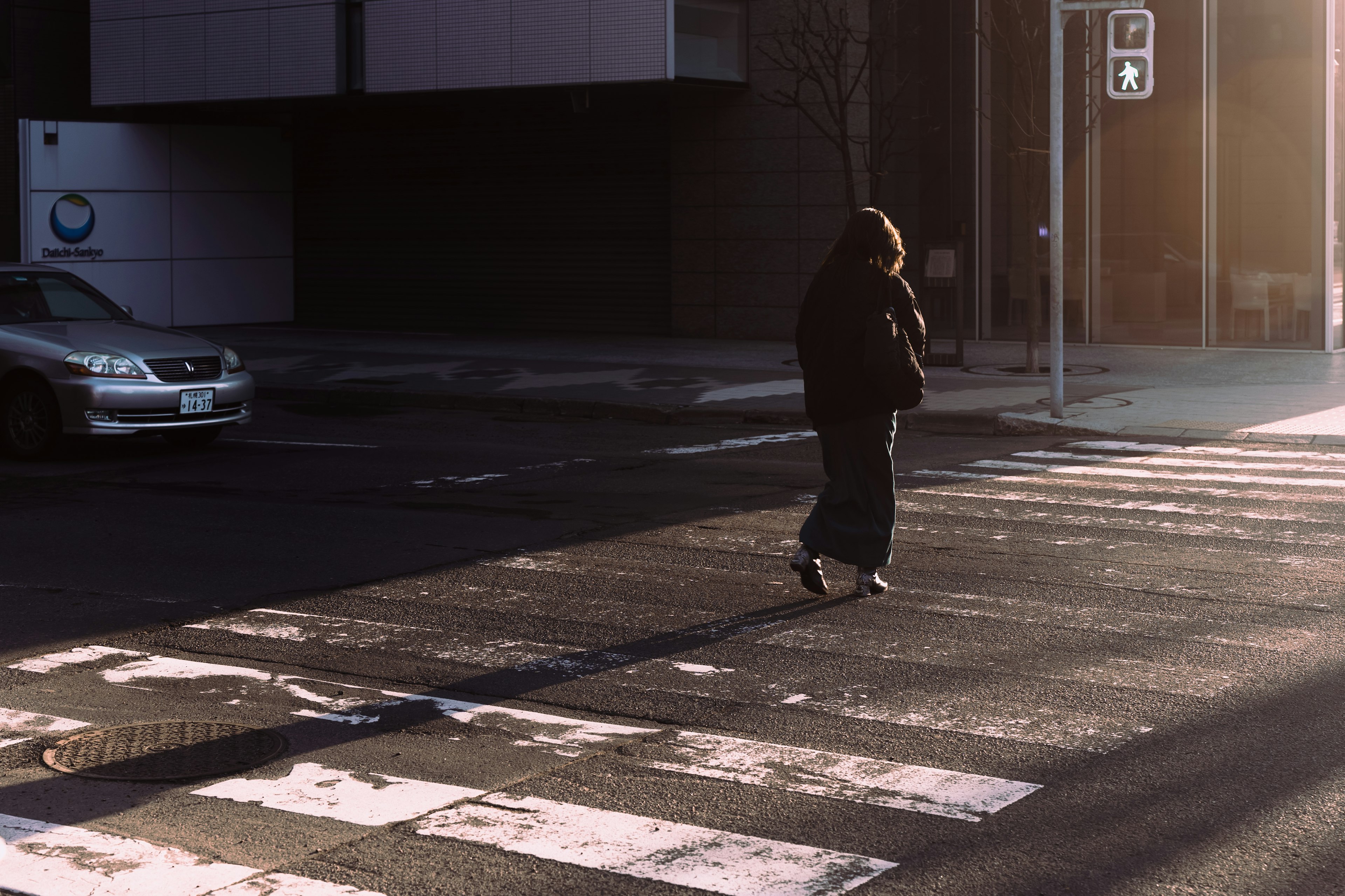 Una persona in un cappotto nero che attraversa la strada con lunghe ombre al sole