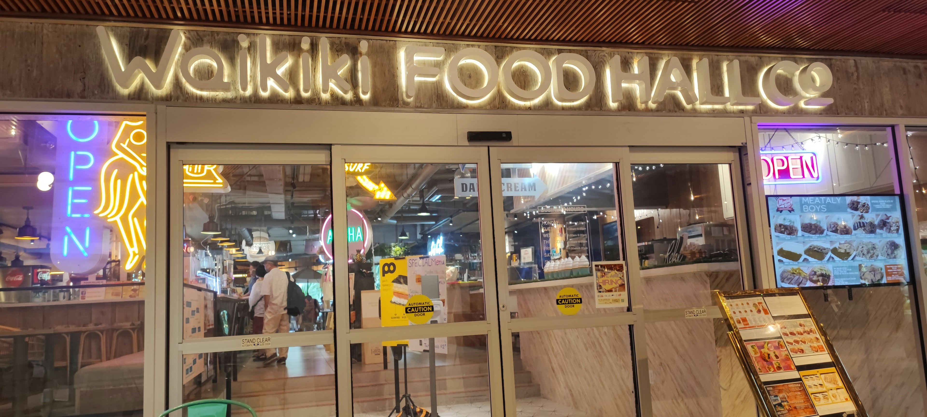 ทางเข้า Waikiki Food Hall พร้อมป้ายไฟนีออนและป้ายเปิดที่มองเห็นได้