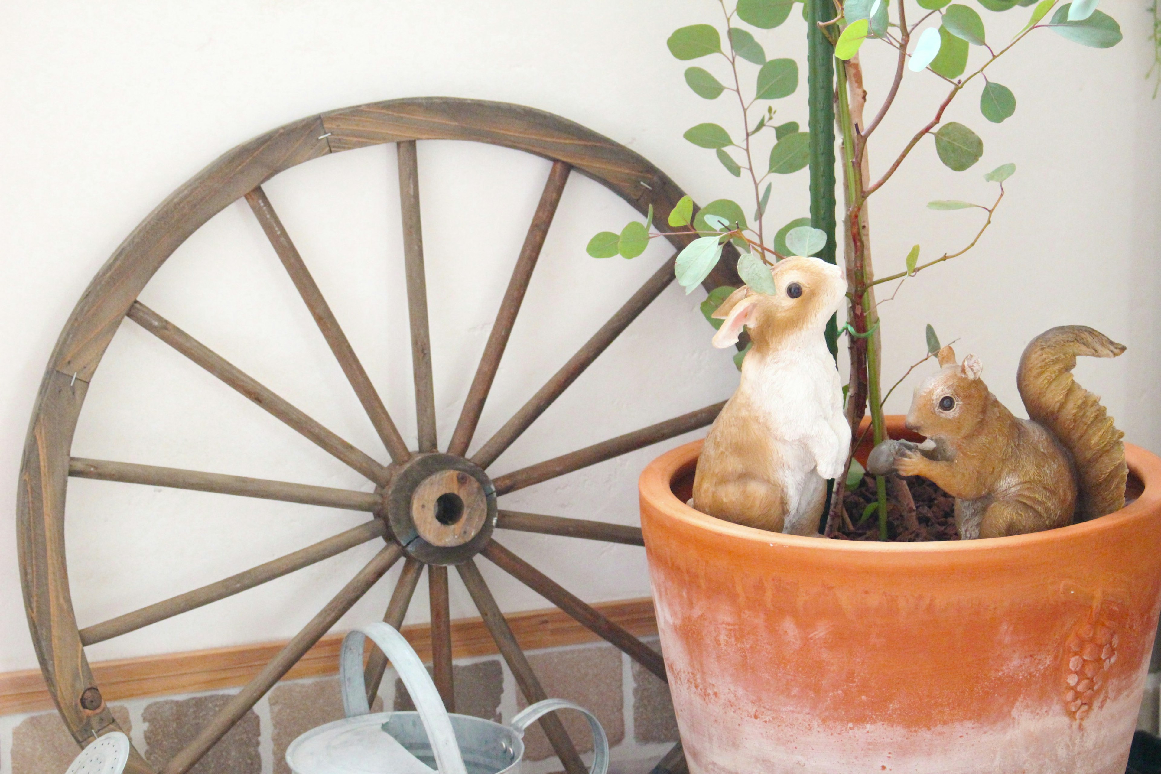 Maceta de cerámica con una planta junto a figuras de conejo y ardilla y una rueda de madera