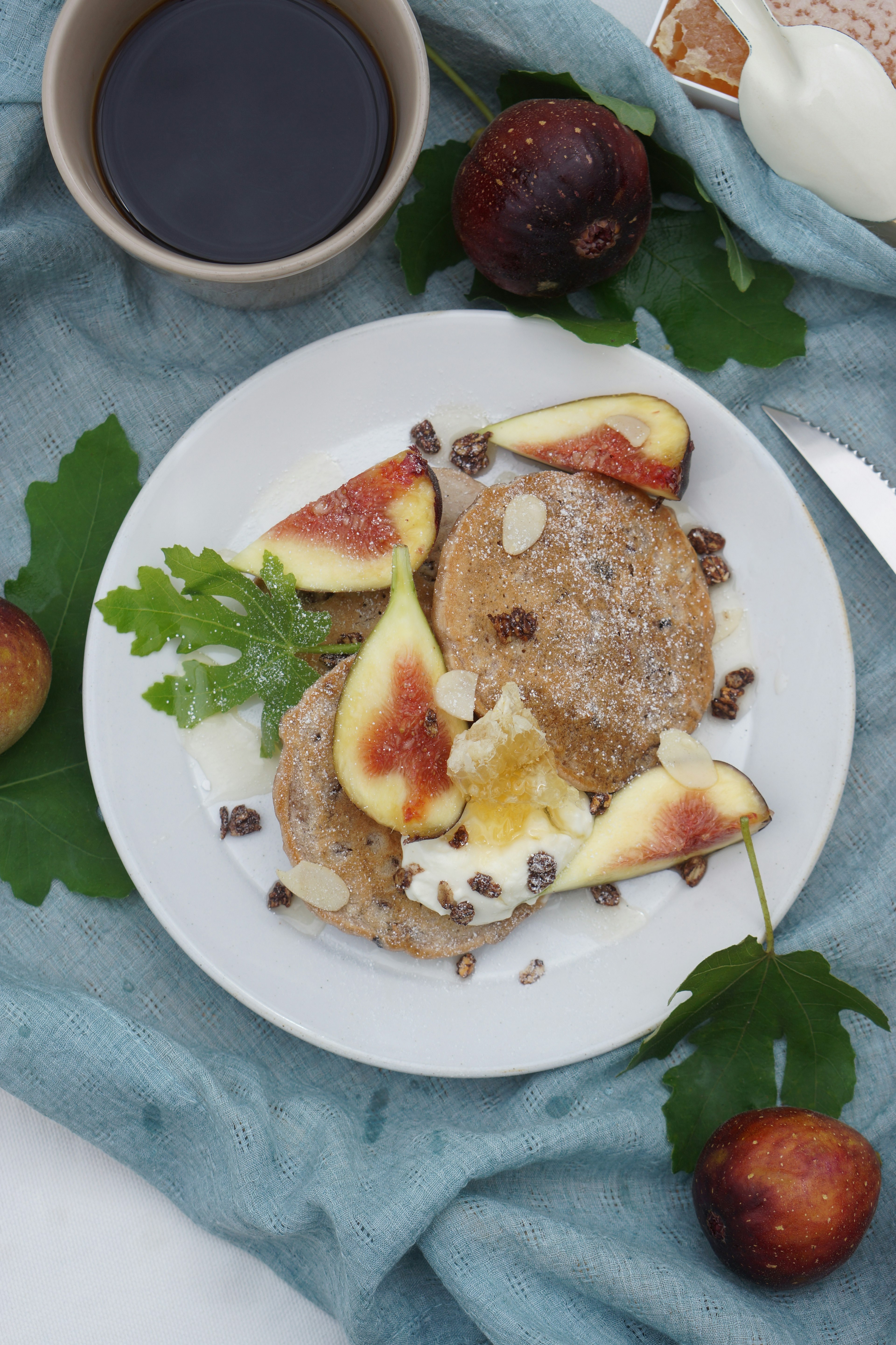 Một đĩa bánh pancake phủ hạt và quả sung trên vải màu xanh với một tách cà phê