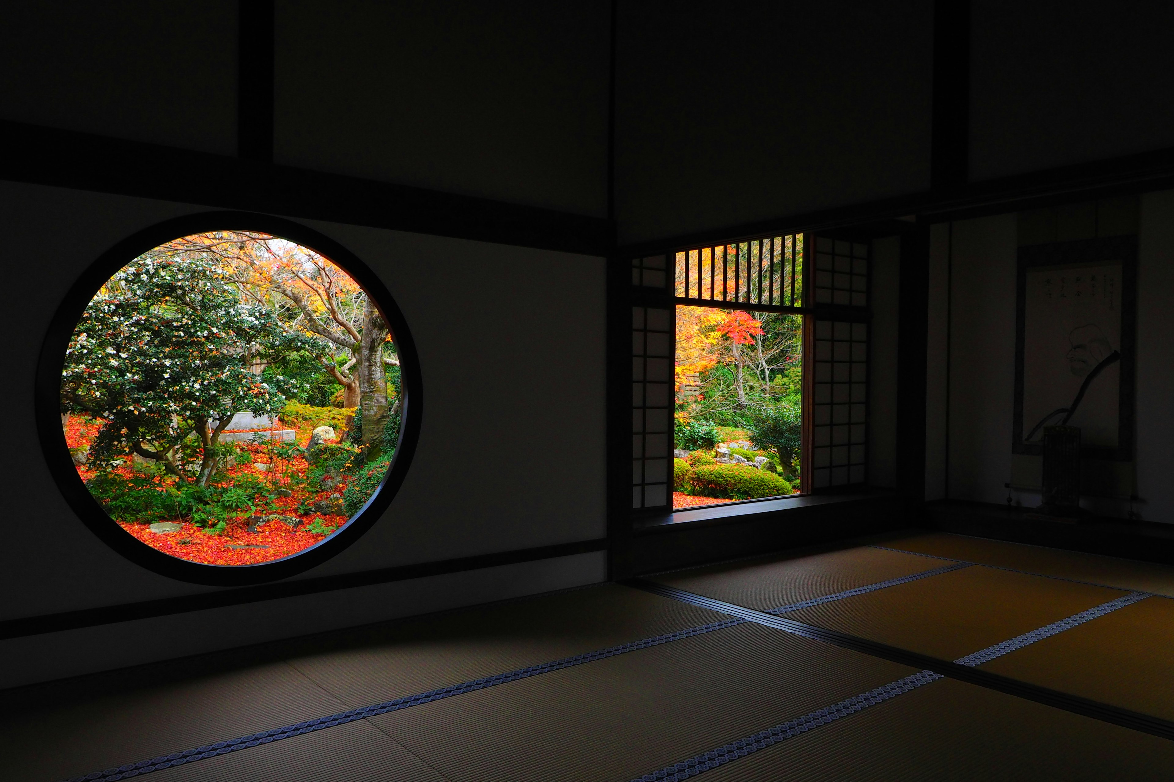 A traditional Japanese room with a circular window showcasing autumn foliage and a garden through shoji