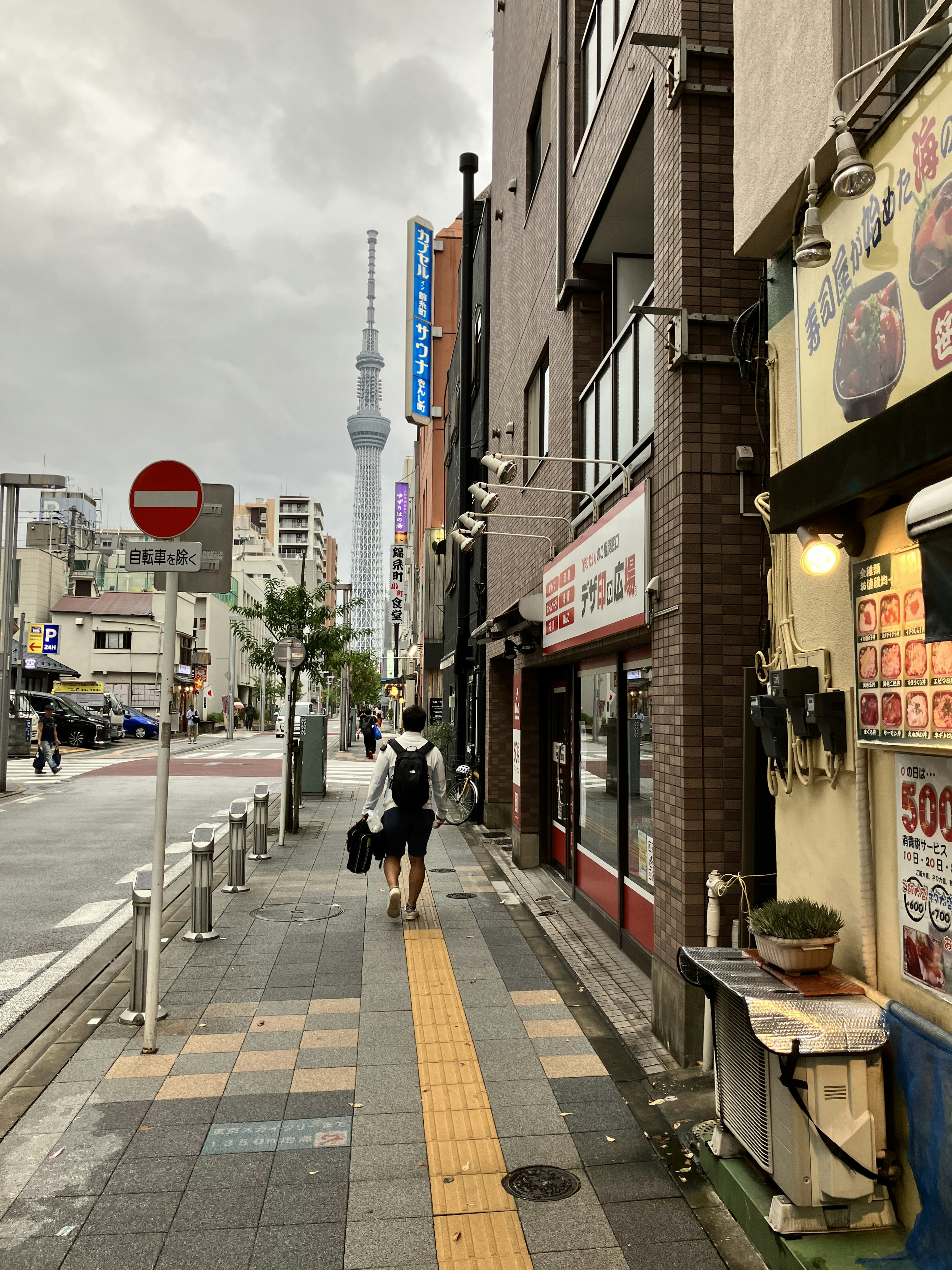 Seseorang yang berjalan di sepanjang jalan dengan Tokyo Skytree terlihat di latar belakang di bawah langit mendung