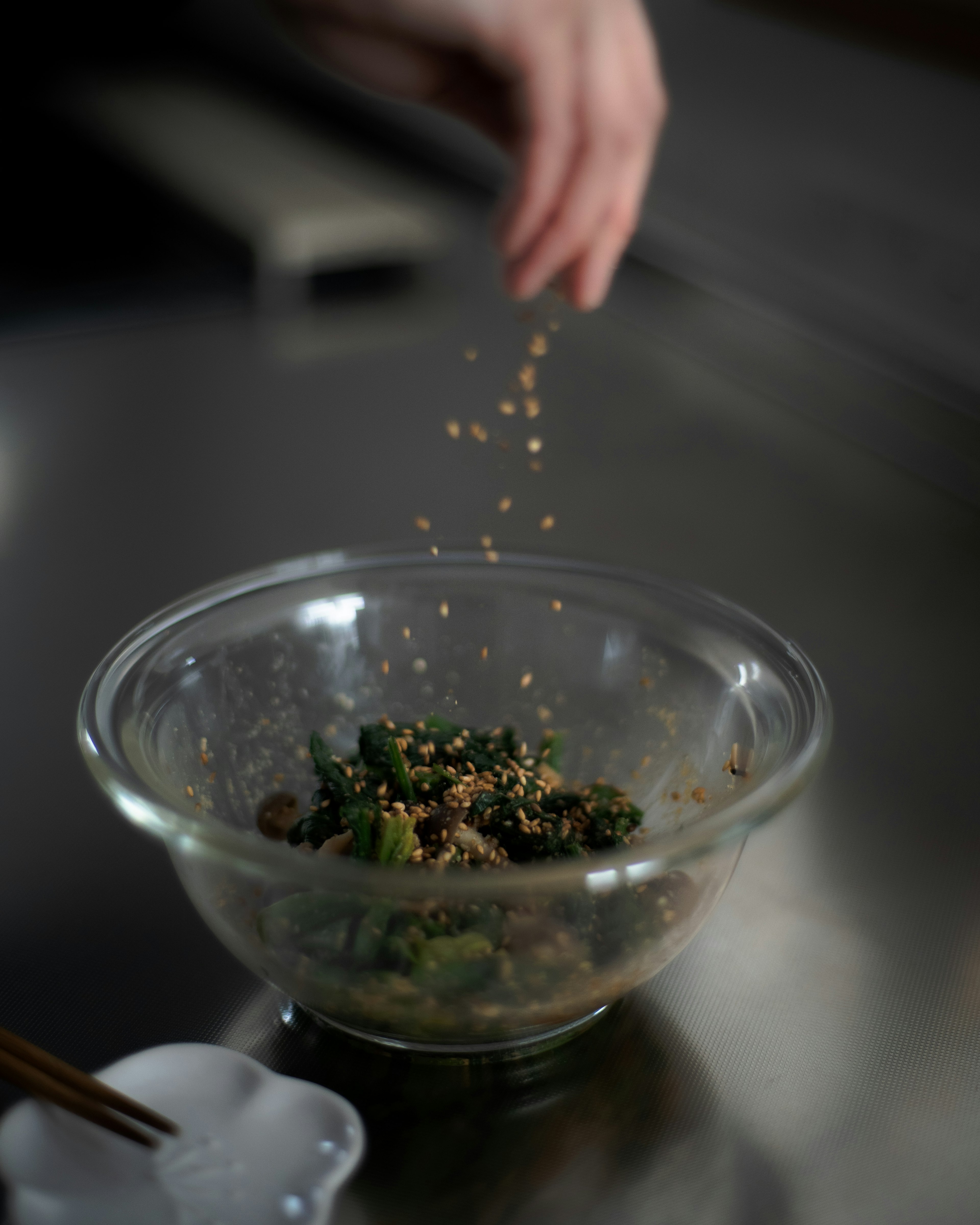 Hand sprinkling sesame seeds over a glass bowl of vegetables