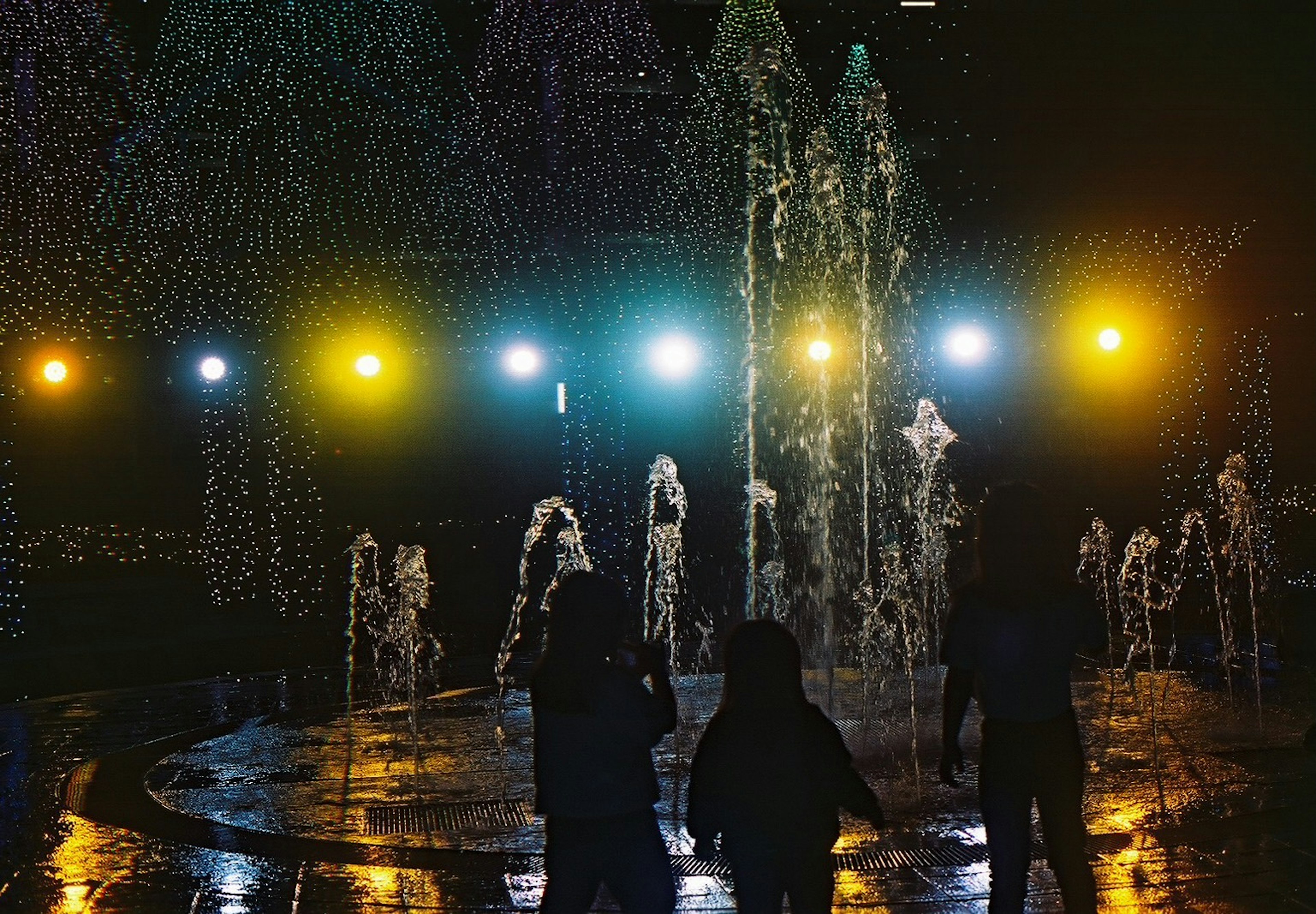 Siluetas de personas frente a una fuente iluminada por la noche