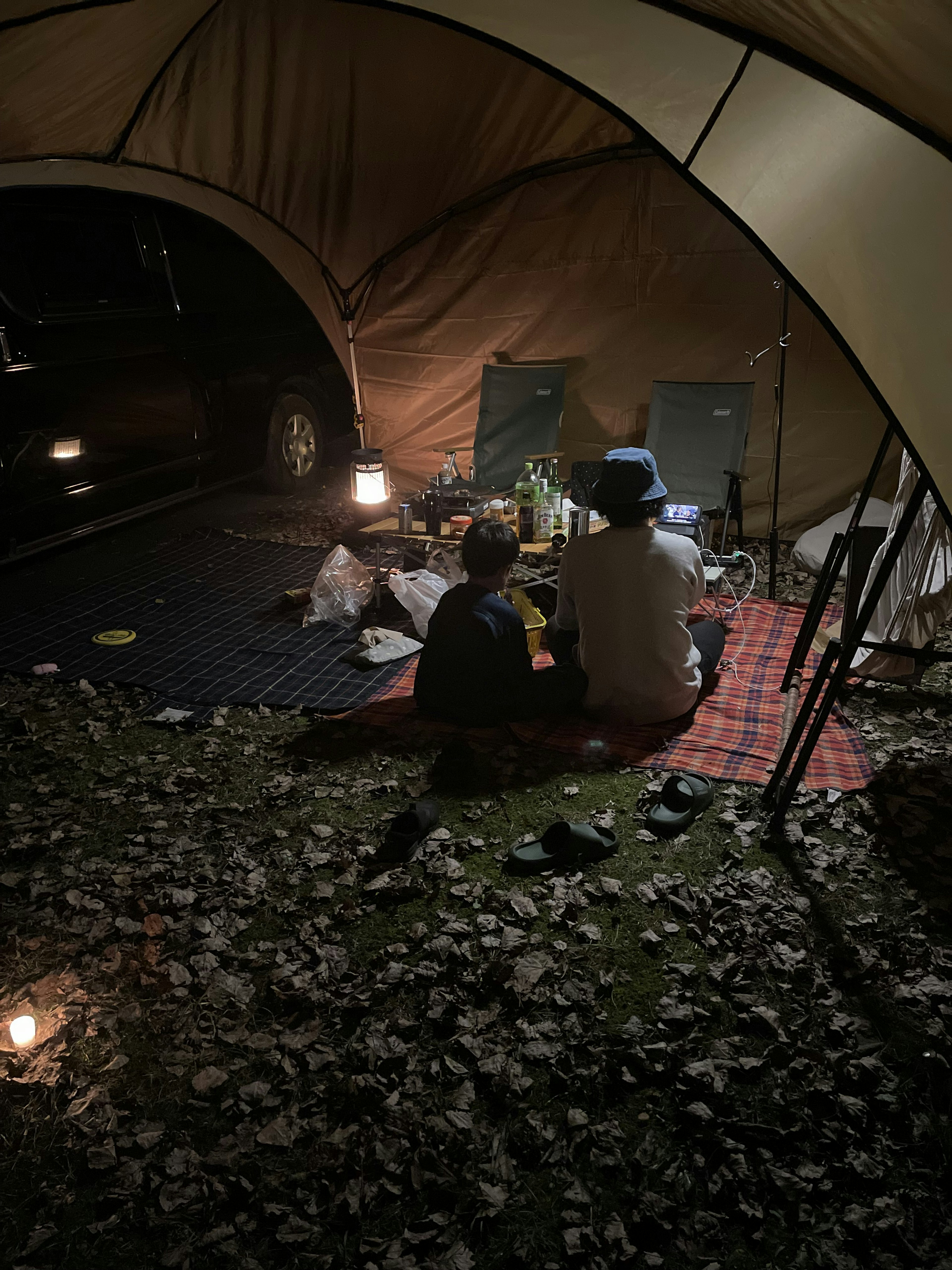 Deux enfants assis à l'intérieur d'une tente de camping la nuit entourés de feuilles et de lanternes