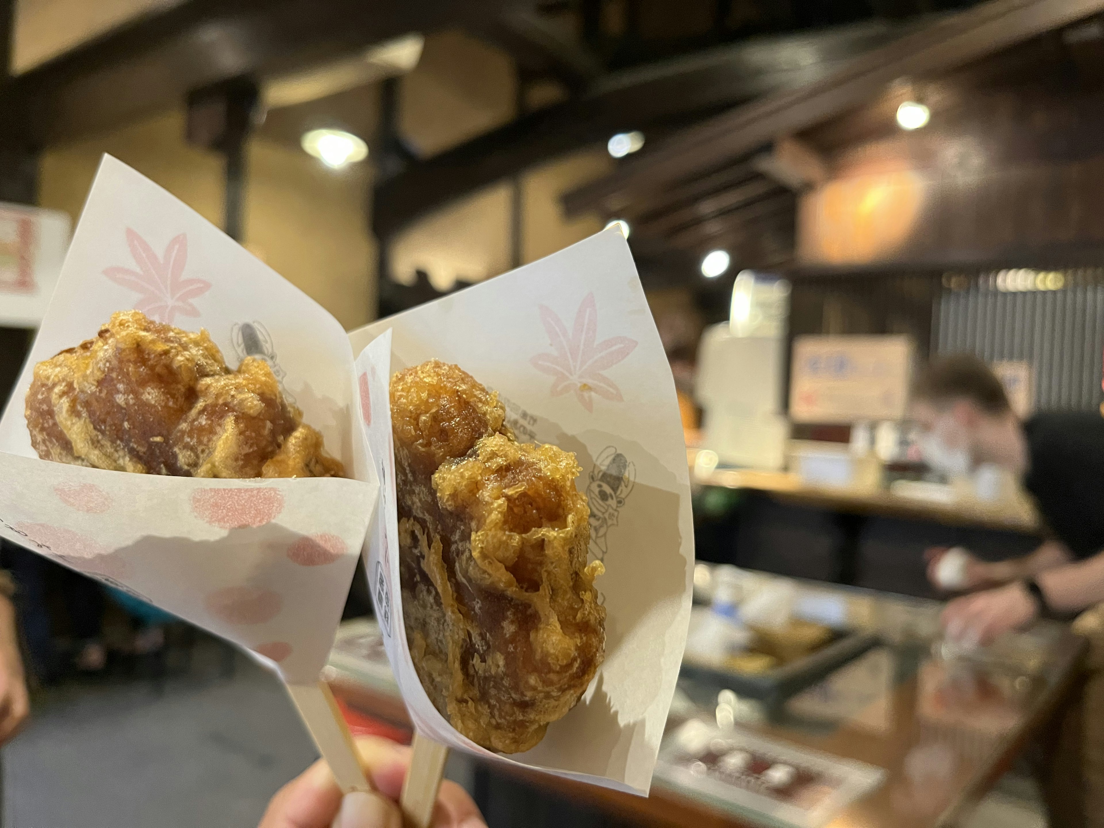 Dua potong ayam goreng dipegang di tangan dibungkus kertas tradisional dengan latar belakang restoran