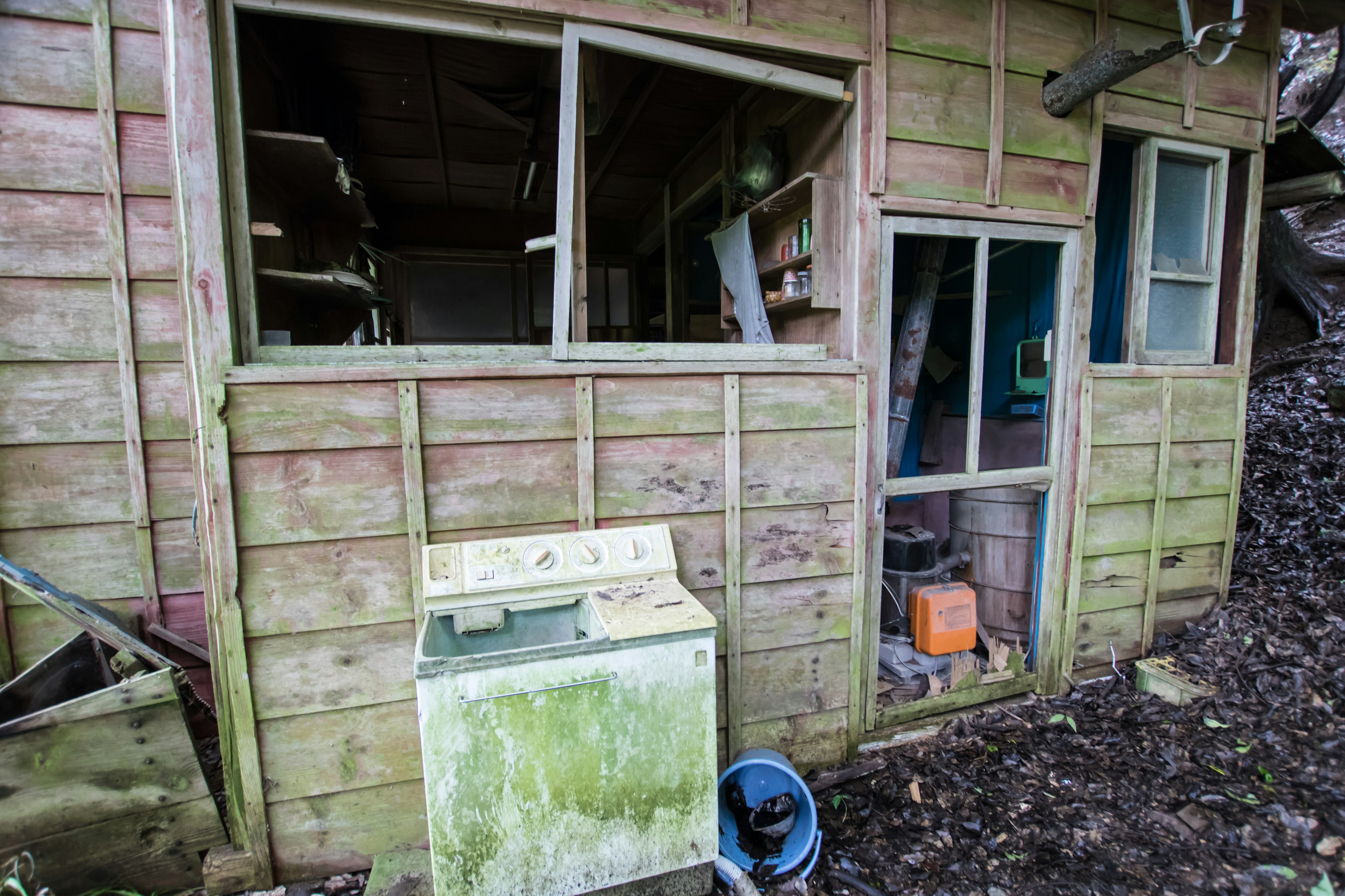 Exterior of an old wooden shed with broken windows green washing machine and surrounding debris