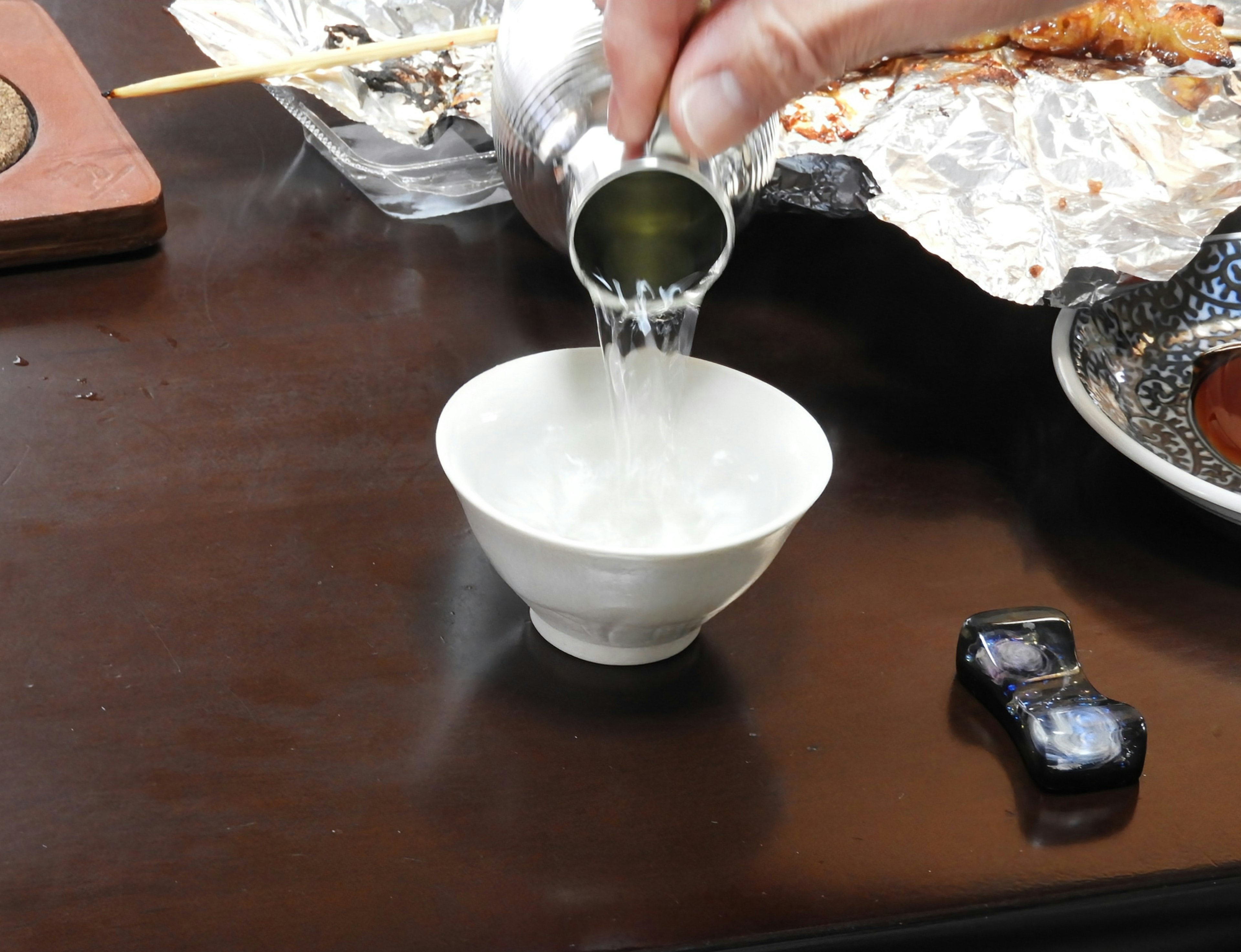 Eine Hand gießt Sake aus einer silbernen Flasche in eine weiße Schüssel auf einem Holztisch mit Gerichten