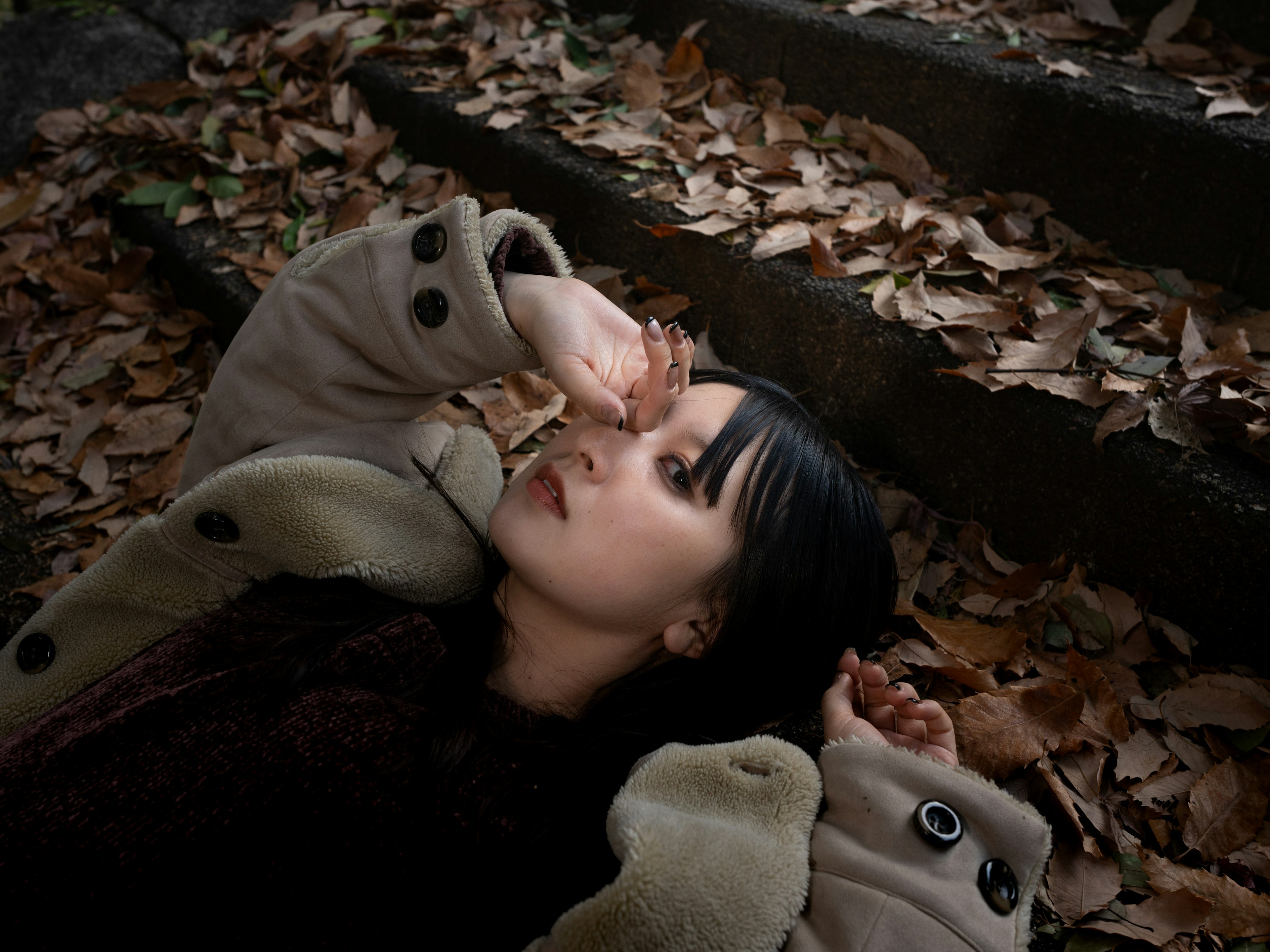 A woman lying on fallen leaves with black hair wearing a coat in a serene natural setting
