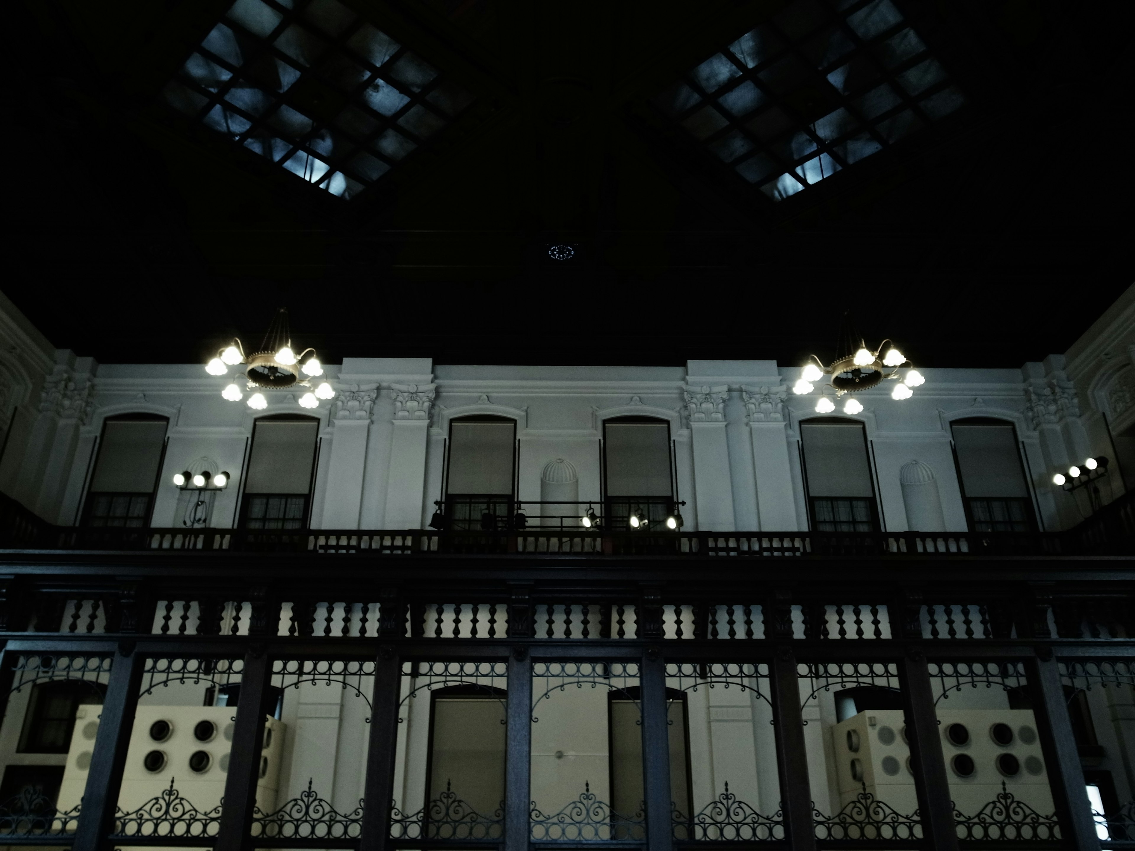 Interior de un edificio histórico con ambiente oscuro, paredes blancas, grandes ventanas y candelabros