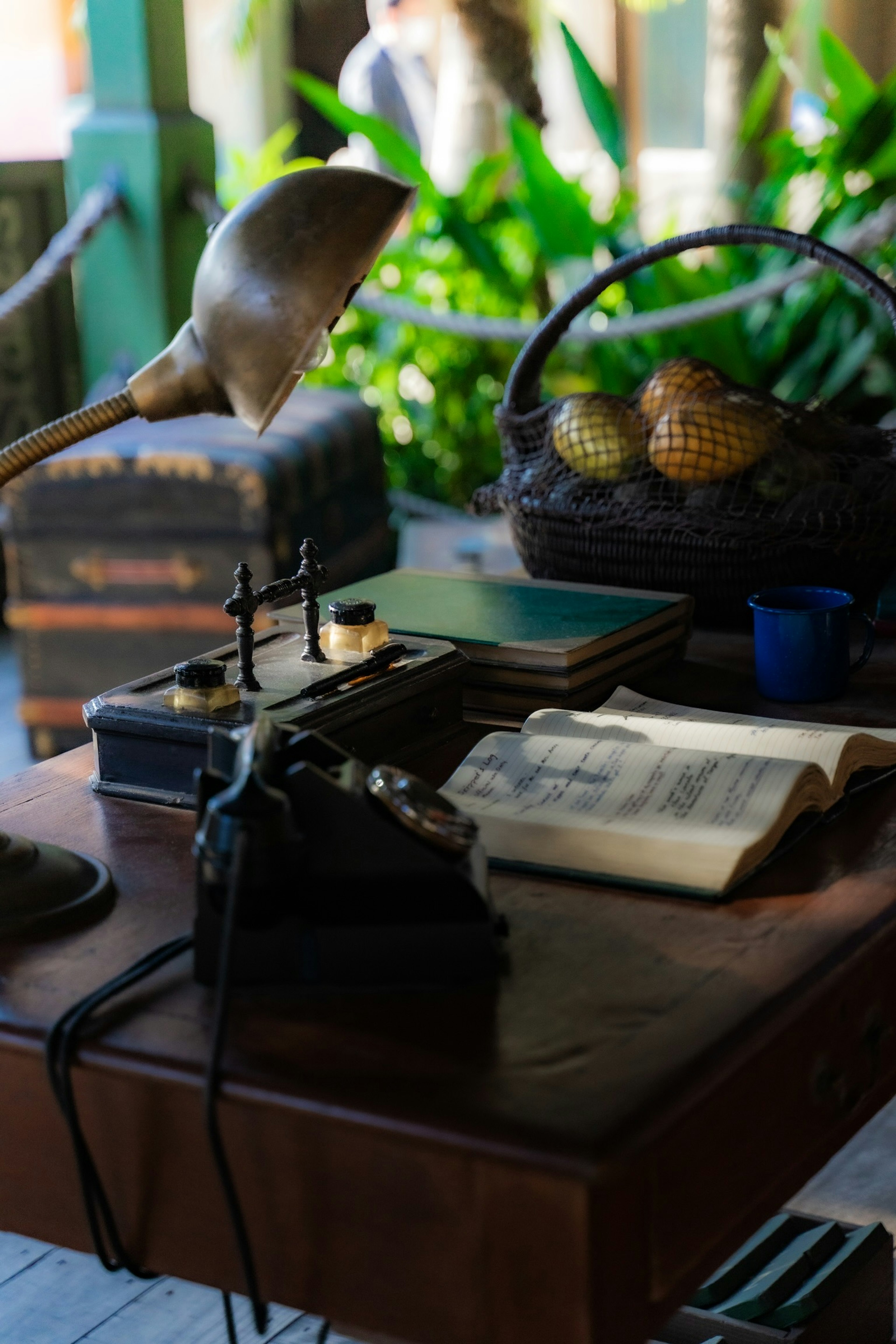 An antique typewriter and a book on a wooden desk with a lamp and a basket of fruits in the background