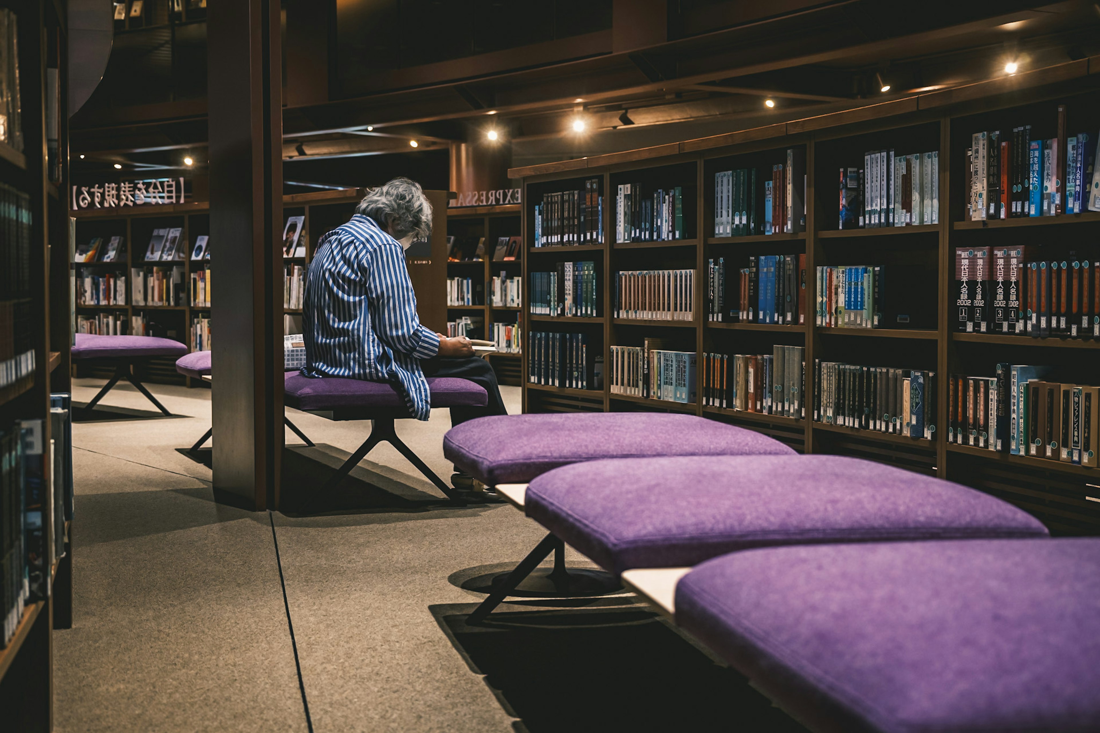 Une personne lisant dans une bibliothèque avec des bancs violets et des étagères de livres